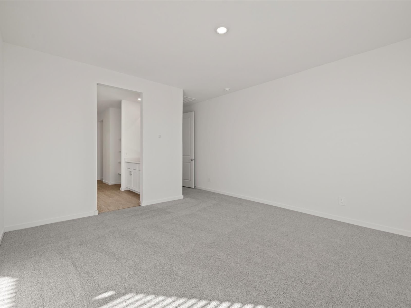 Empty room with light gray carpet, white walls, a single ceiling light, and an open doorway leading to a bathroom with wood-look flooring.