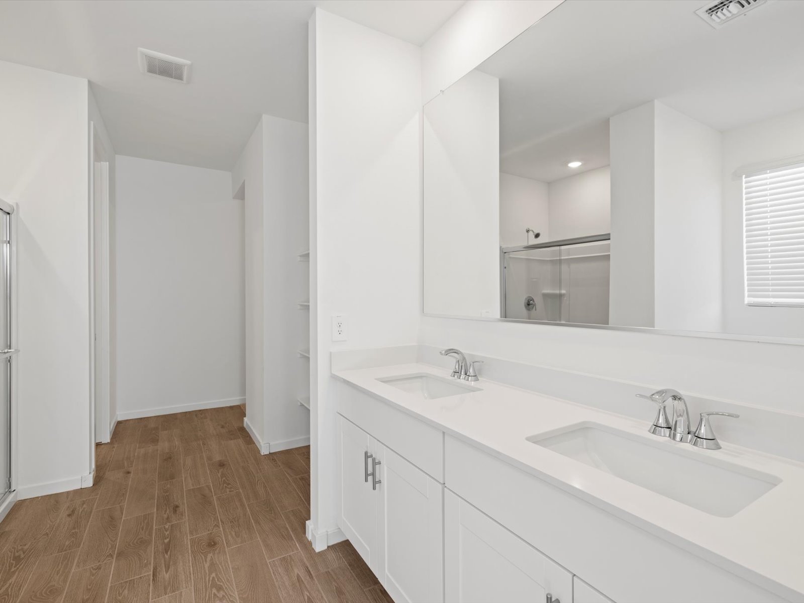 Modern bathroom with double sinks, white cabinets, large mirror, wood flooring, and a walk-in shower with glass doors. Natural light enters through a window with blinds.