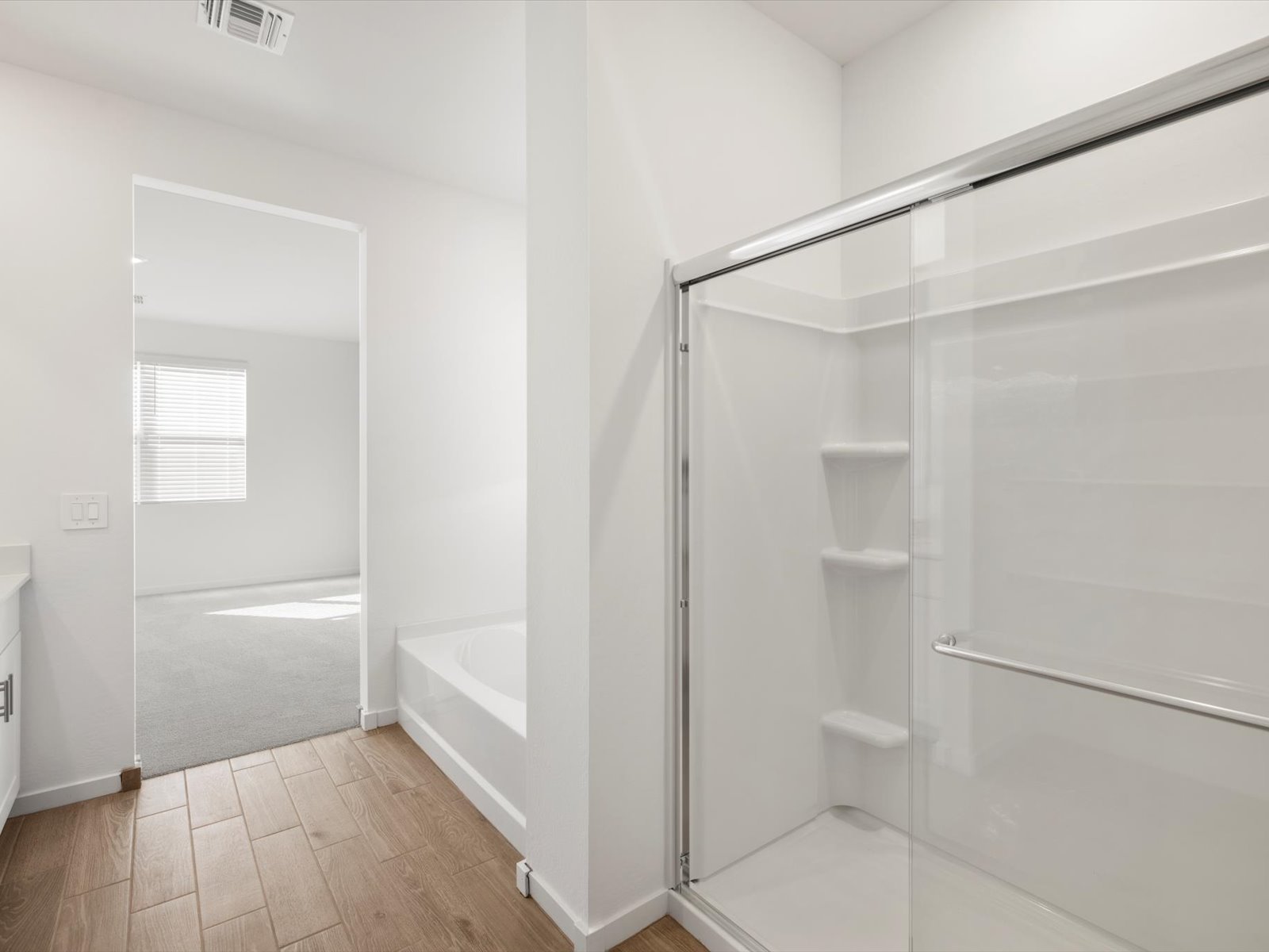 Modern bathroom with a glass shower, built-in shelves, a bathtub, wood-look flooring, and a view into a bright, carpeted bedroom.