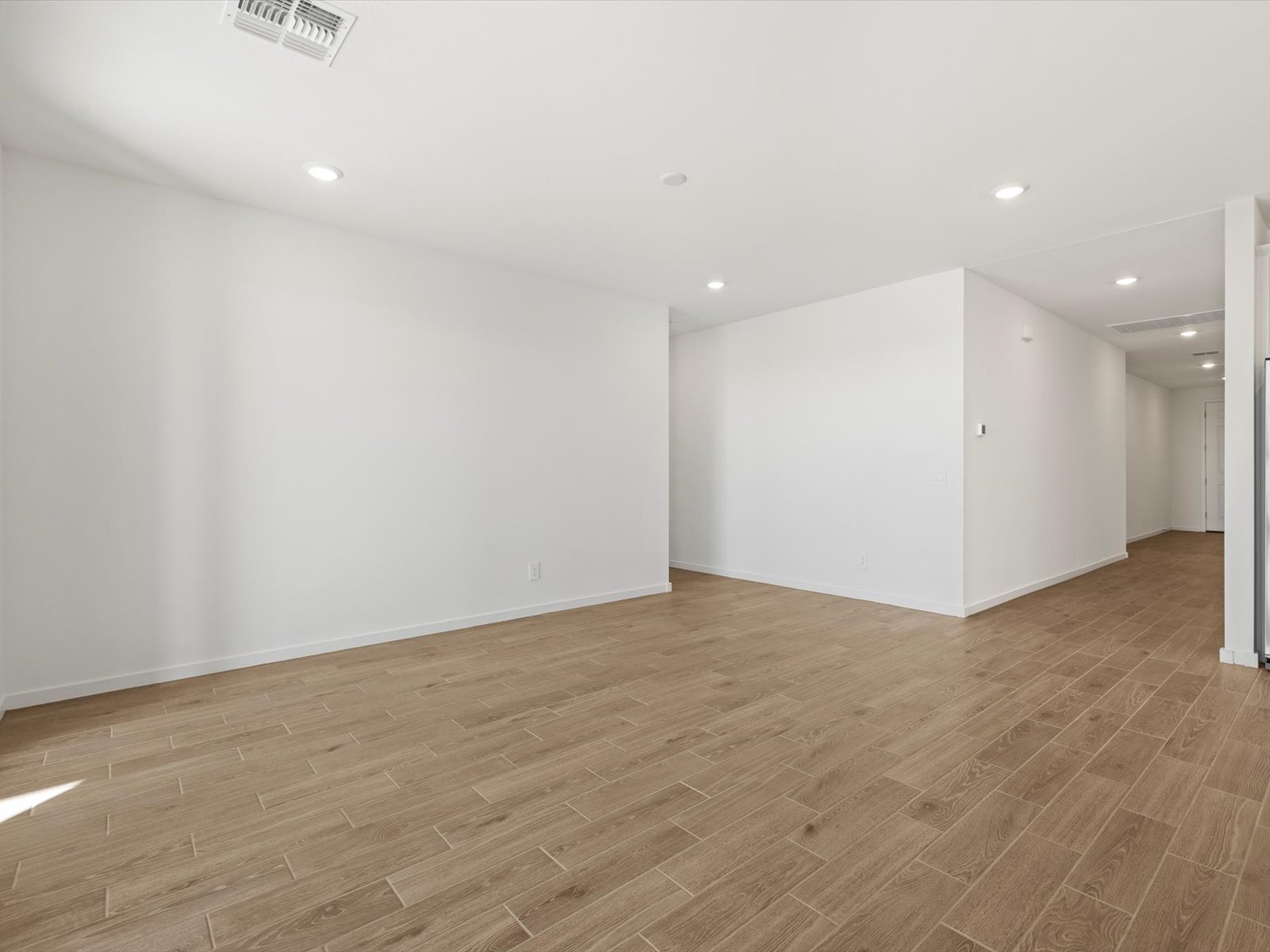 Empty room with white walls, recessed ceiling lights, wood-look tile flooring, and a hallway leading to other areas of the house.