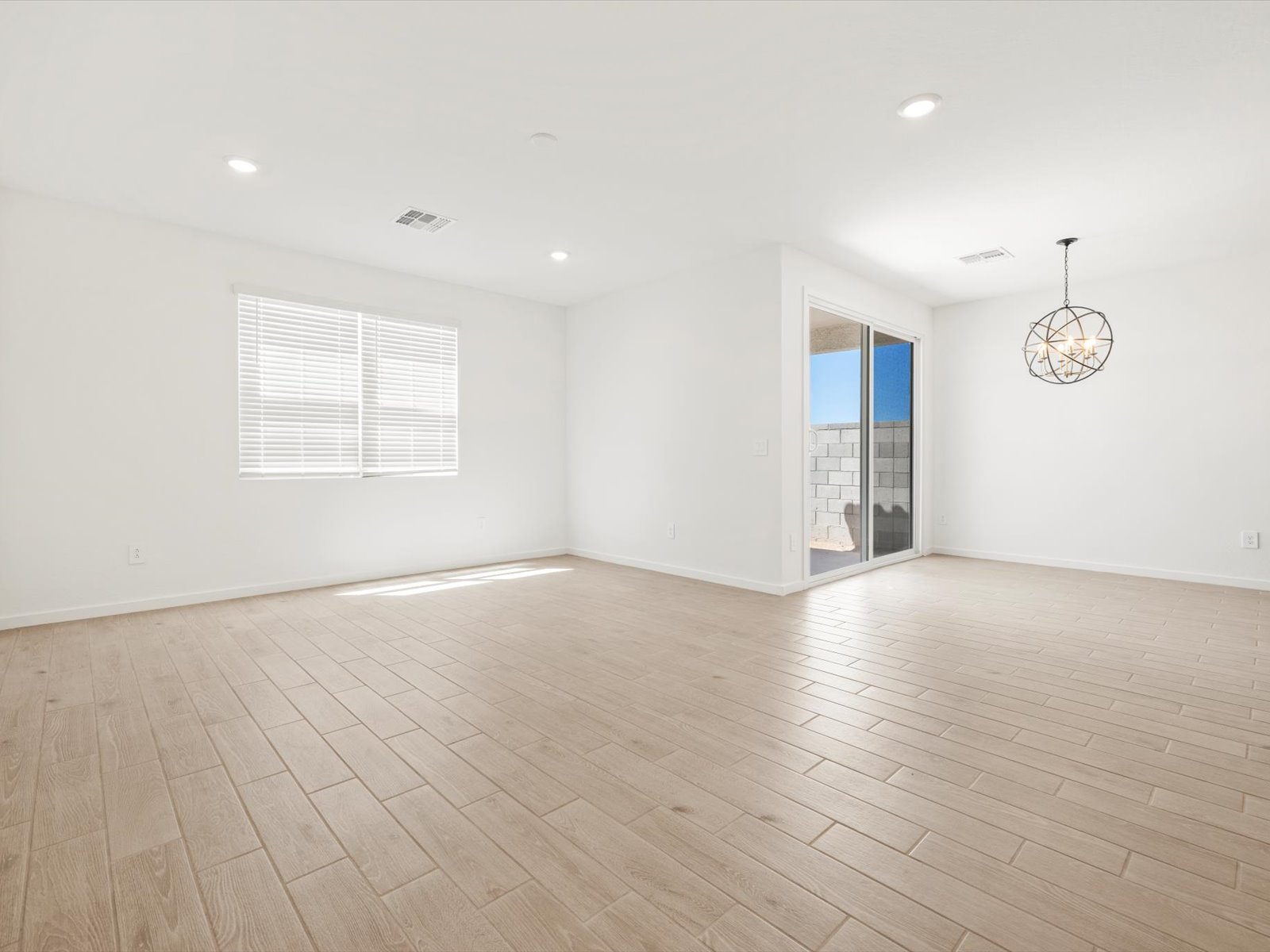 Empty, unfurnished room with light wood tile flooring, white walls, a window, recessed ceiling lights, and a sliding glass door leading to a small patio with a brick wall outside.