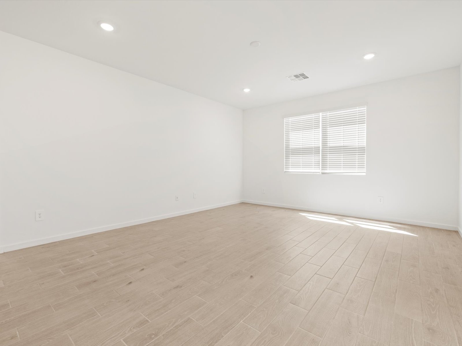Empty room with white walls, light wood tile flooring, recessed ceiling lights, and a window with closed blinds letting in sunlight.