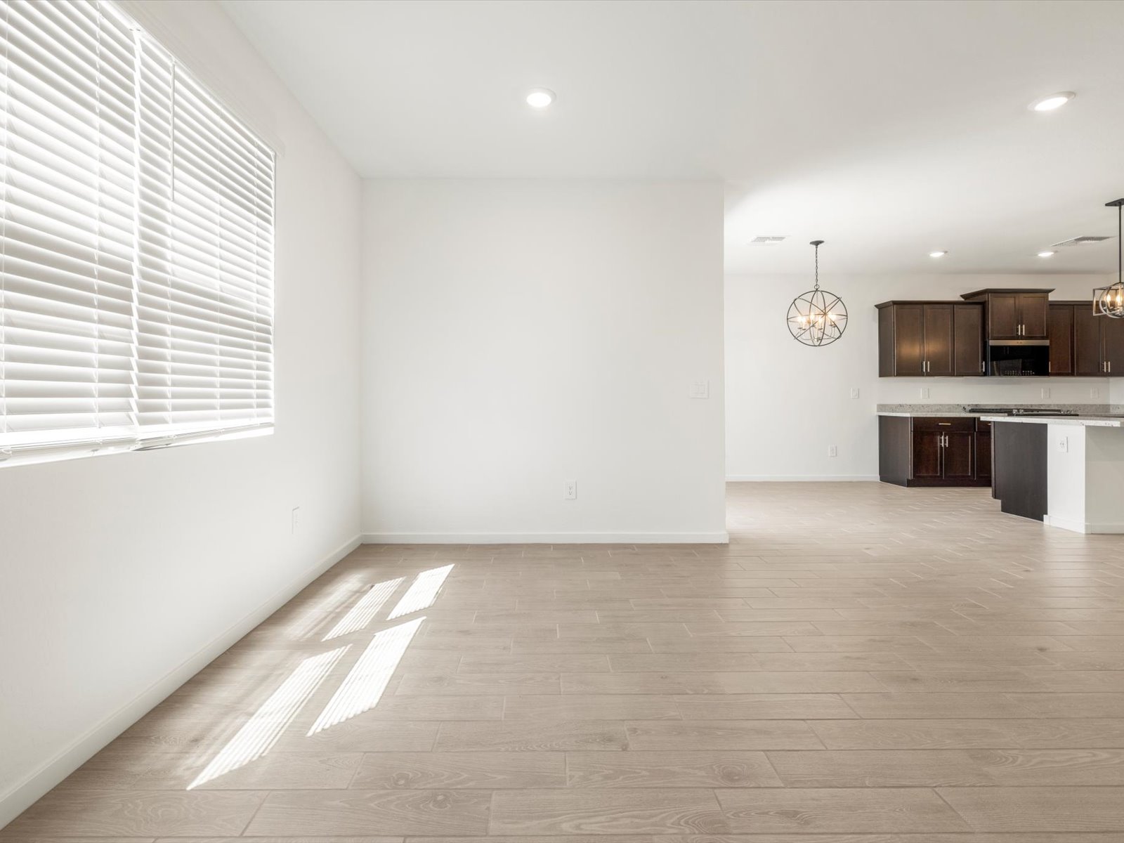 Unfurnished open living space with light wood flooring, white walls, large window with blinds, and a view of a modern kitchen with dark cabinets and pendant lights.