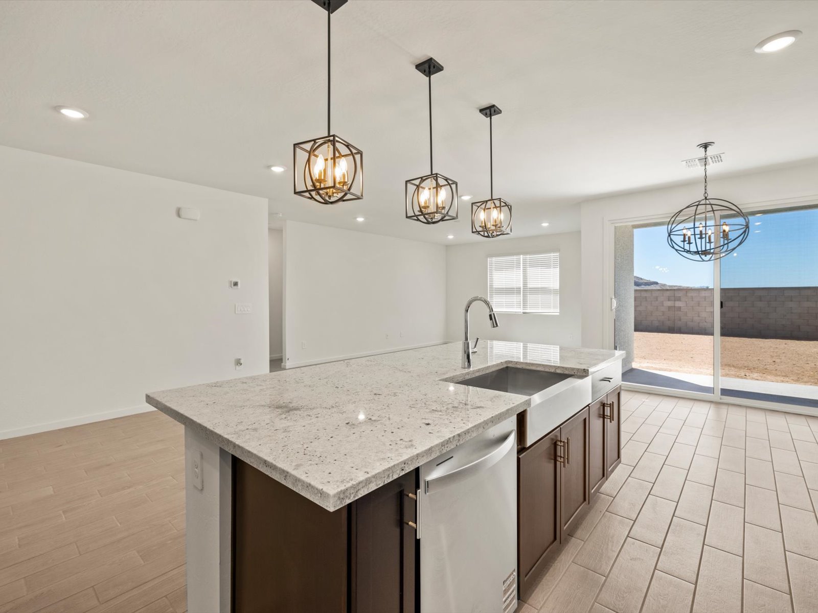Modern kitchen with a large island, pendant lights, stainless steel sink, and dishwasher, adjacent to a bright open living area with sliding glass doors to a fenced backyard.
