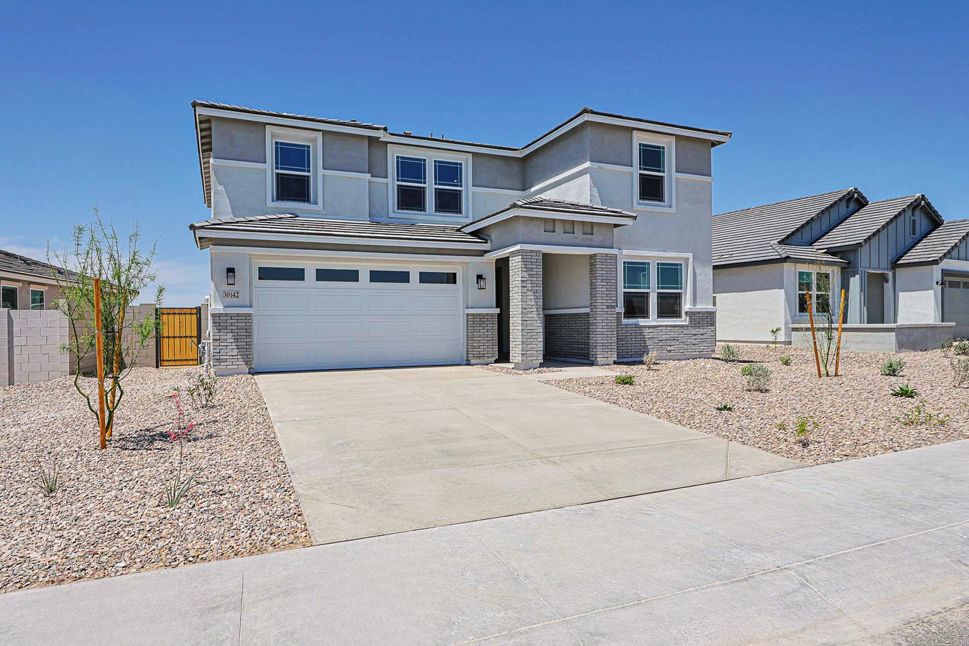 Modern two-story house with a double garage, gray trim, and a rock-covered front yard, located in a suburban neighborhood under a clear blue sky.