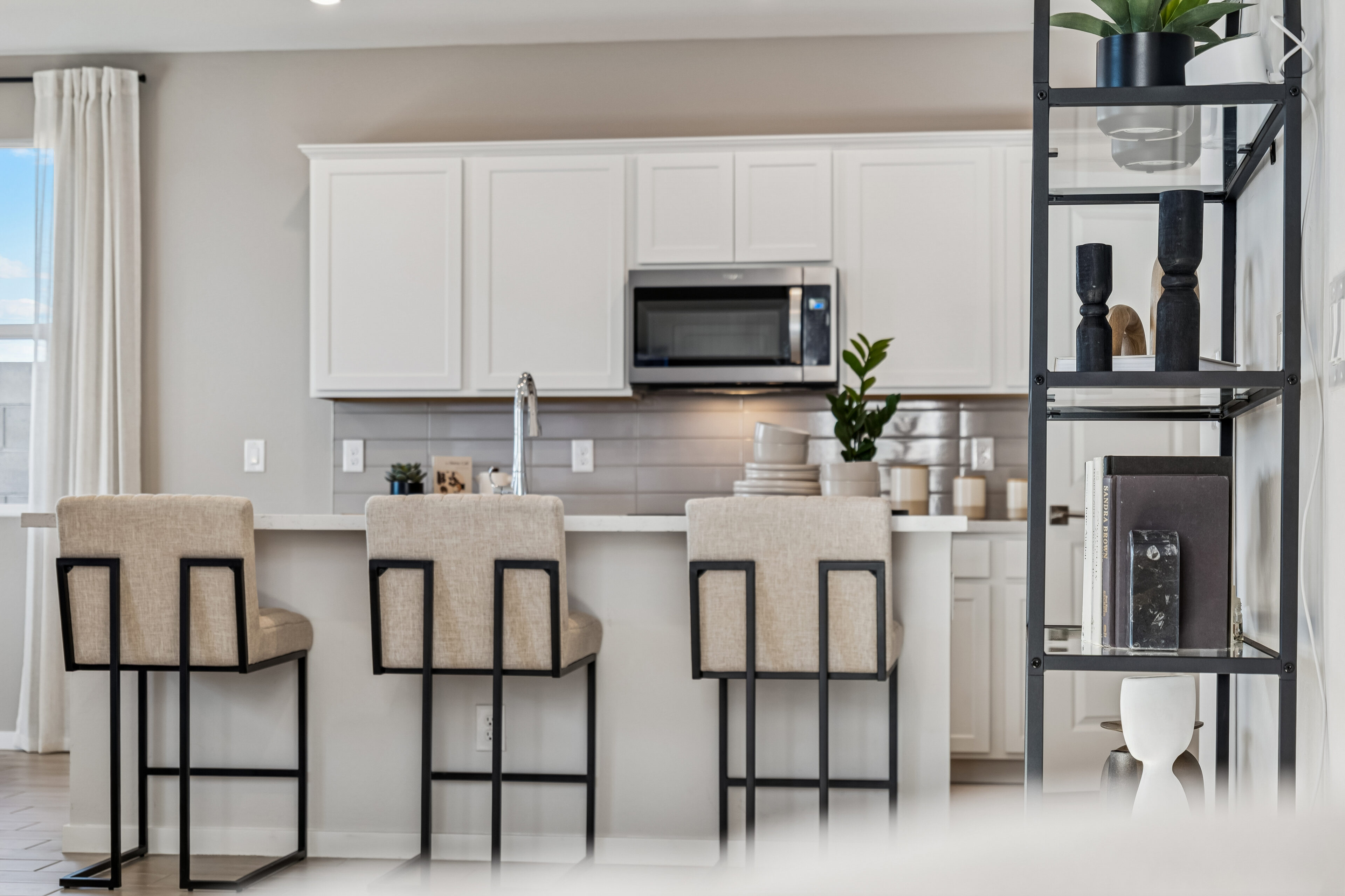 Modern kitchen with white cabinets, a countertop with three beige barstools, a stainless steel microwave, and a black metal shelf with decorative items.
