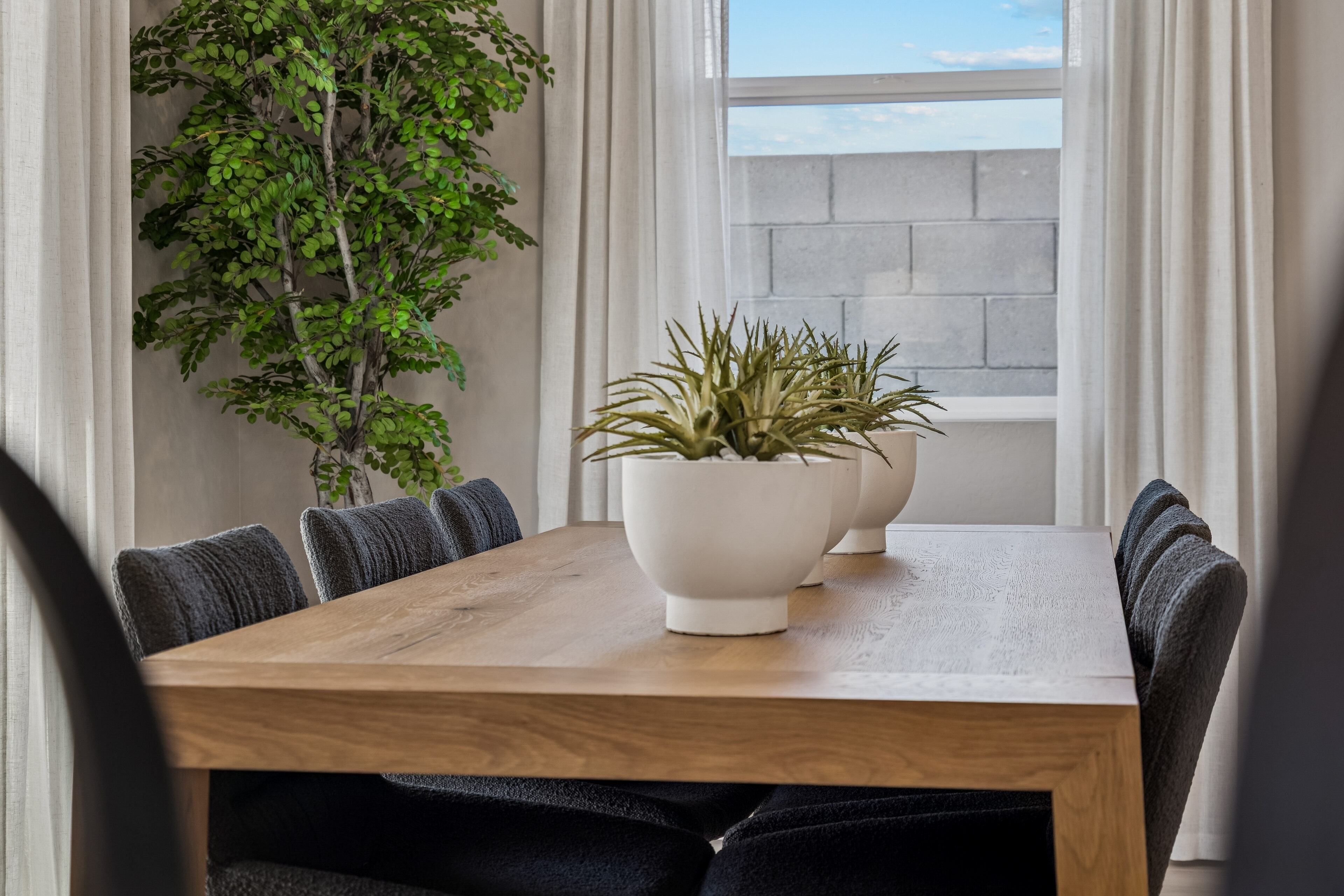 A wooden dining table with six dark chairs and two potted plants sits by a window with white curtains; a green plant is in the corner.