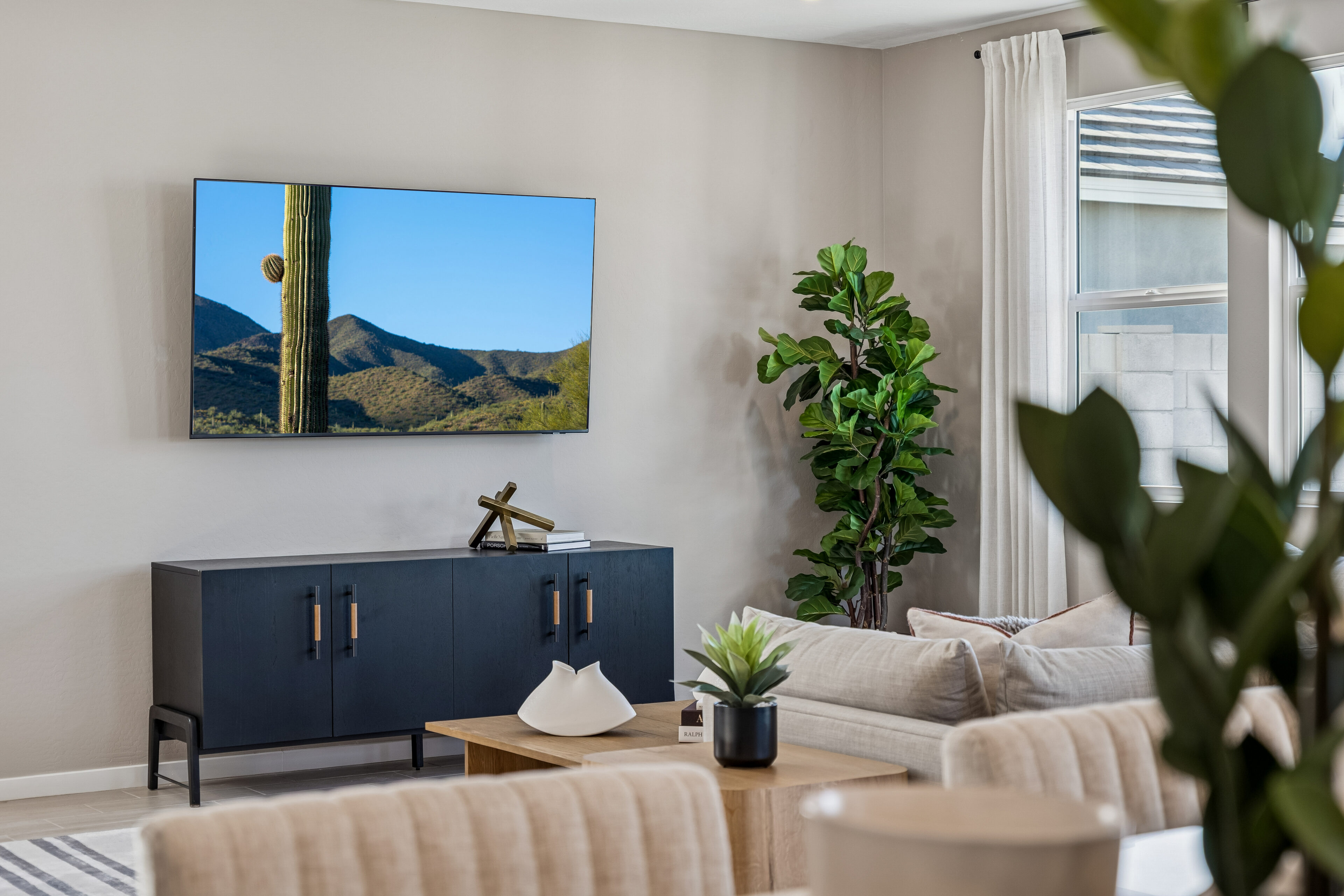 Modern living room with a wall-mounted TV displaying a desert scene, a black cabinet below, beige sofa, wooden coffee table, and a potted plant near a window with white curtains.