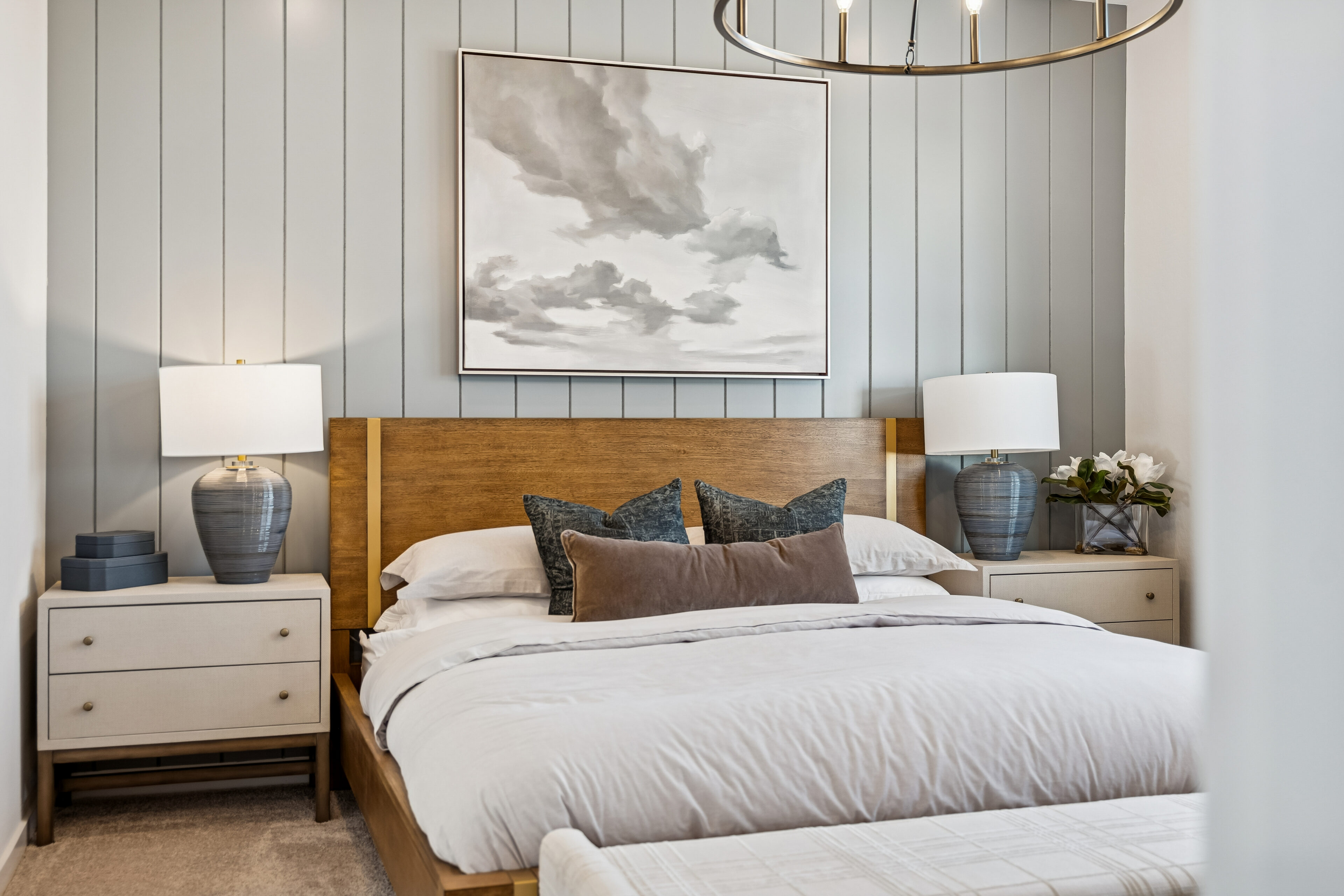 Modern bedroom with a wooden bed frame, white bedding, gray accent pillows, two nightstands with lamps, and a cloud painting hanging above the headboard.