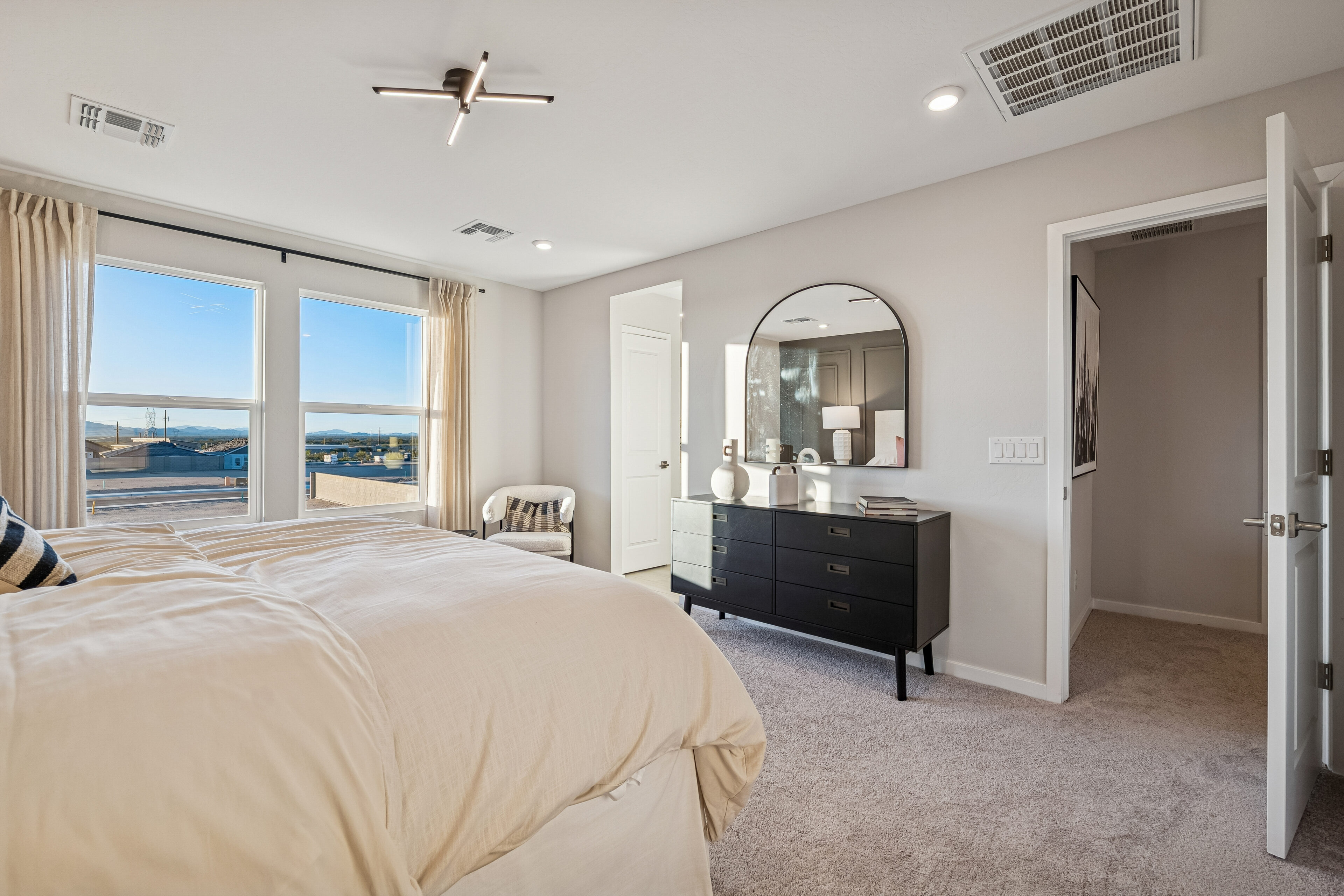 Modern bedroom with beige bedding, black dresser, round mirror, armchair, carpeted floor, large windows, and open doors leading to a hallway and closet.