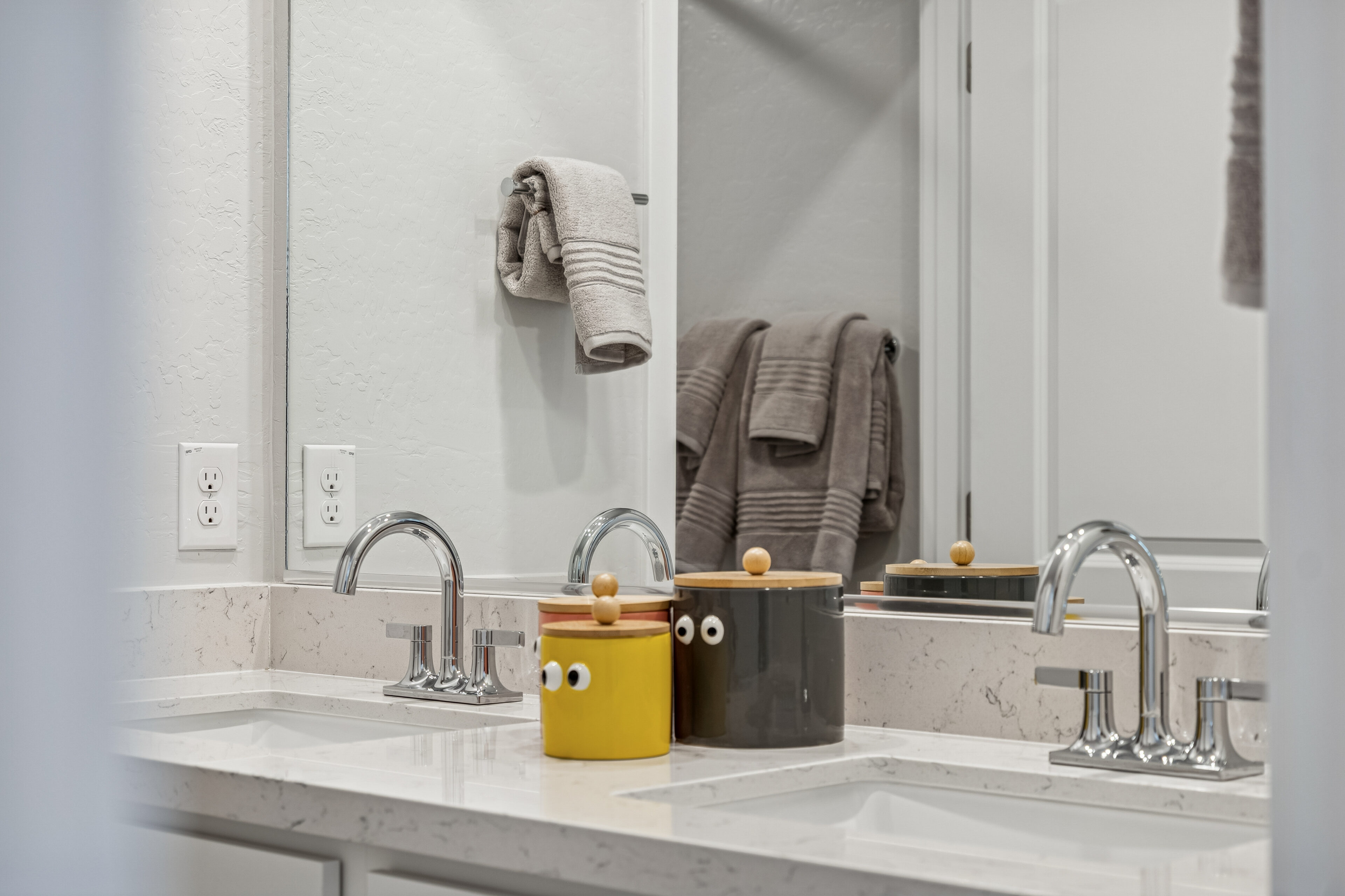 Double bathroom sinks with chrome faucets, two containers with googly eyes on the counter, and folded towels hanging on the wall reflected in a mirror.