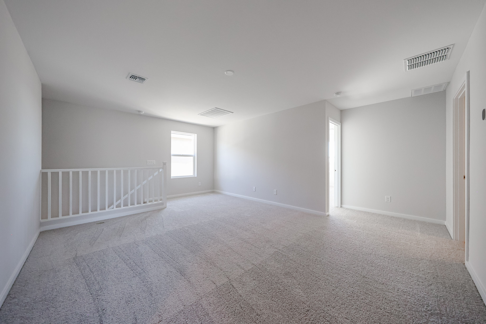 Empty carpeted room with white walls, a window, a doorway, and a staircase with a white railing, all under ceiling vents.