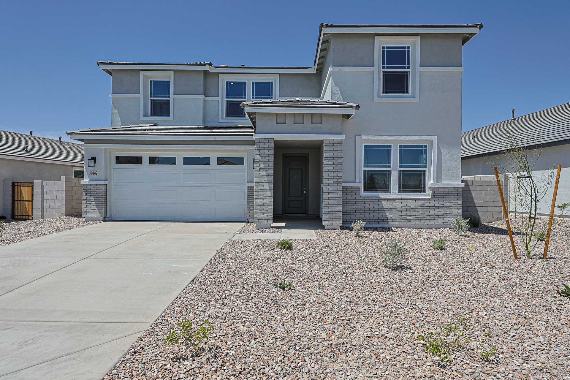 Two-story modern house with a double garage, light gray exterior, and a front yard landscaped with rocks and small plants under a clear blue sky.