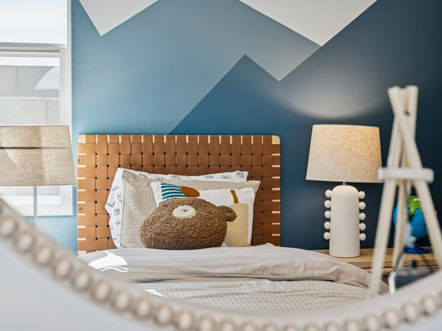 A neatly made bed with a bear pillow, a woven headboard, and a lamp on a nightstand, reflected in a round mirror against a blue and white geometric wall.