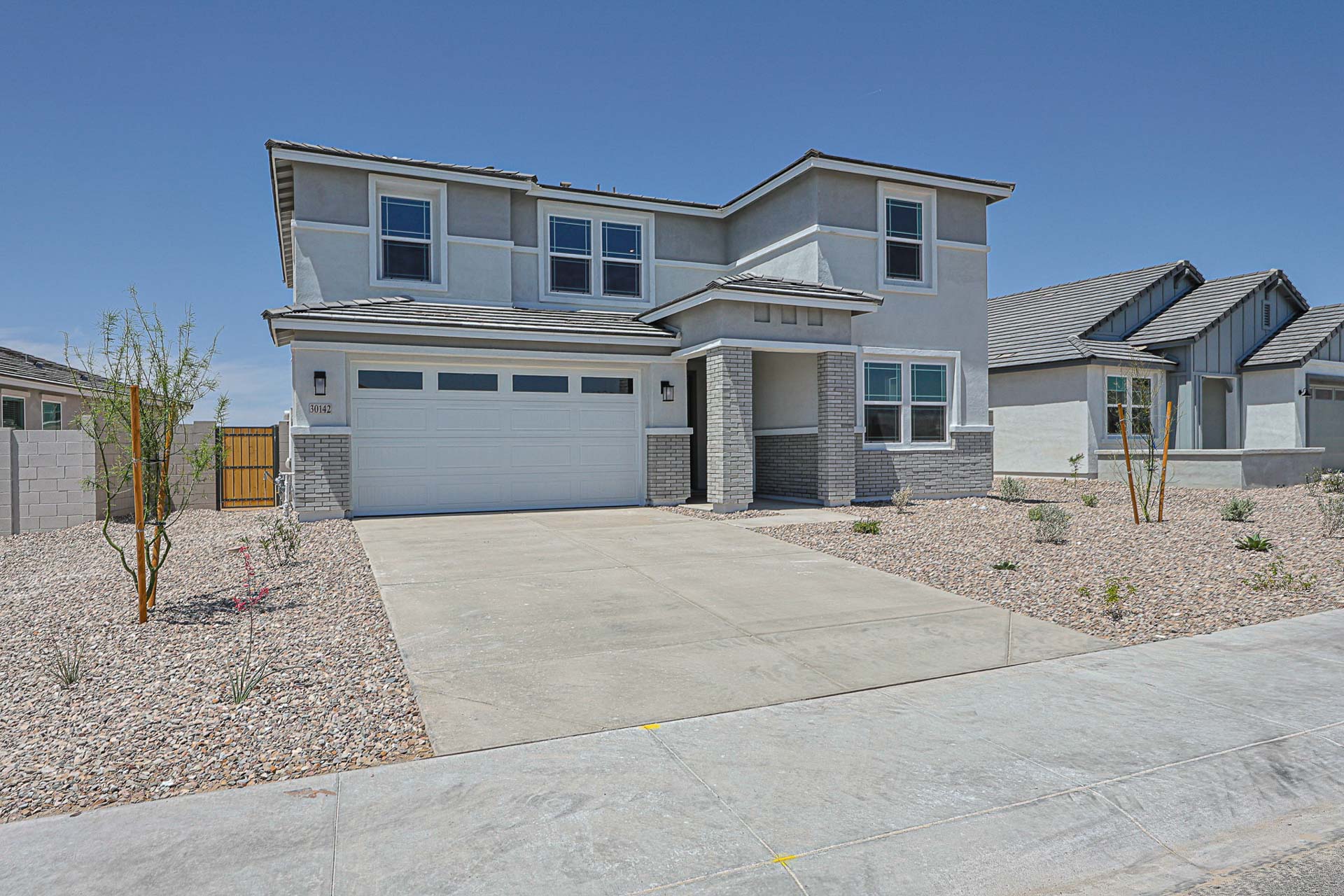 Two-story modern house with a gray exterior, attached two-car garage, and minimal desert landscaping on a sunny day.