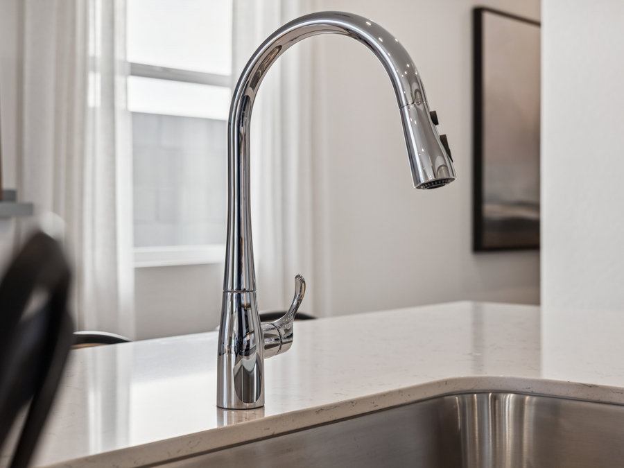 A modern chrome kitchen faucet is mounted on a white countertop next to a stainless steel sink in a bright, contemporary kitchen.