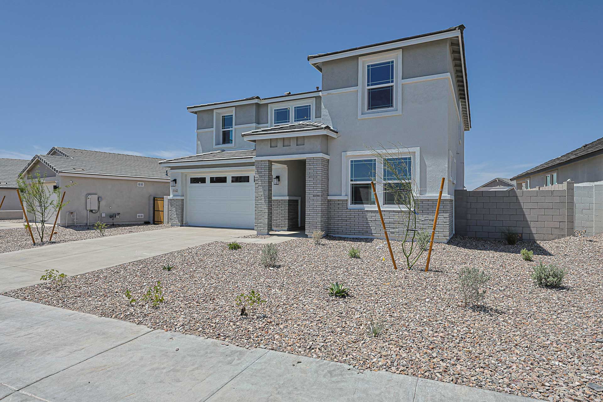 Two-story modern house with a gray exterior, attached garage, and xeriscaped front yard with rocks and sparse plants under a clear blue sky.