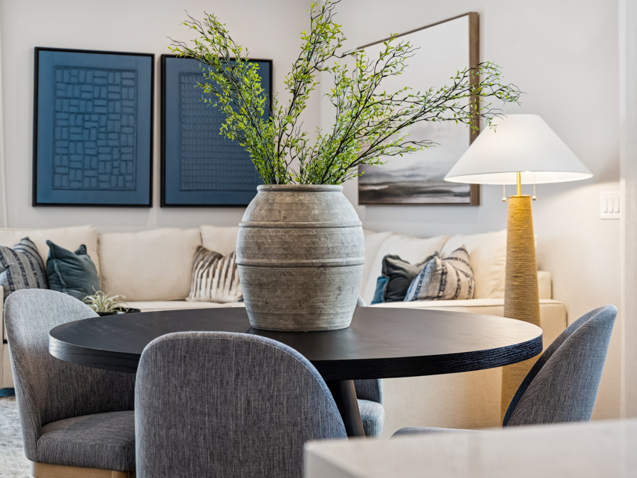 A round black table with a large gray vase of green branches, surrounded by gray chairs, in a modern living room with art and a lamp.