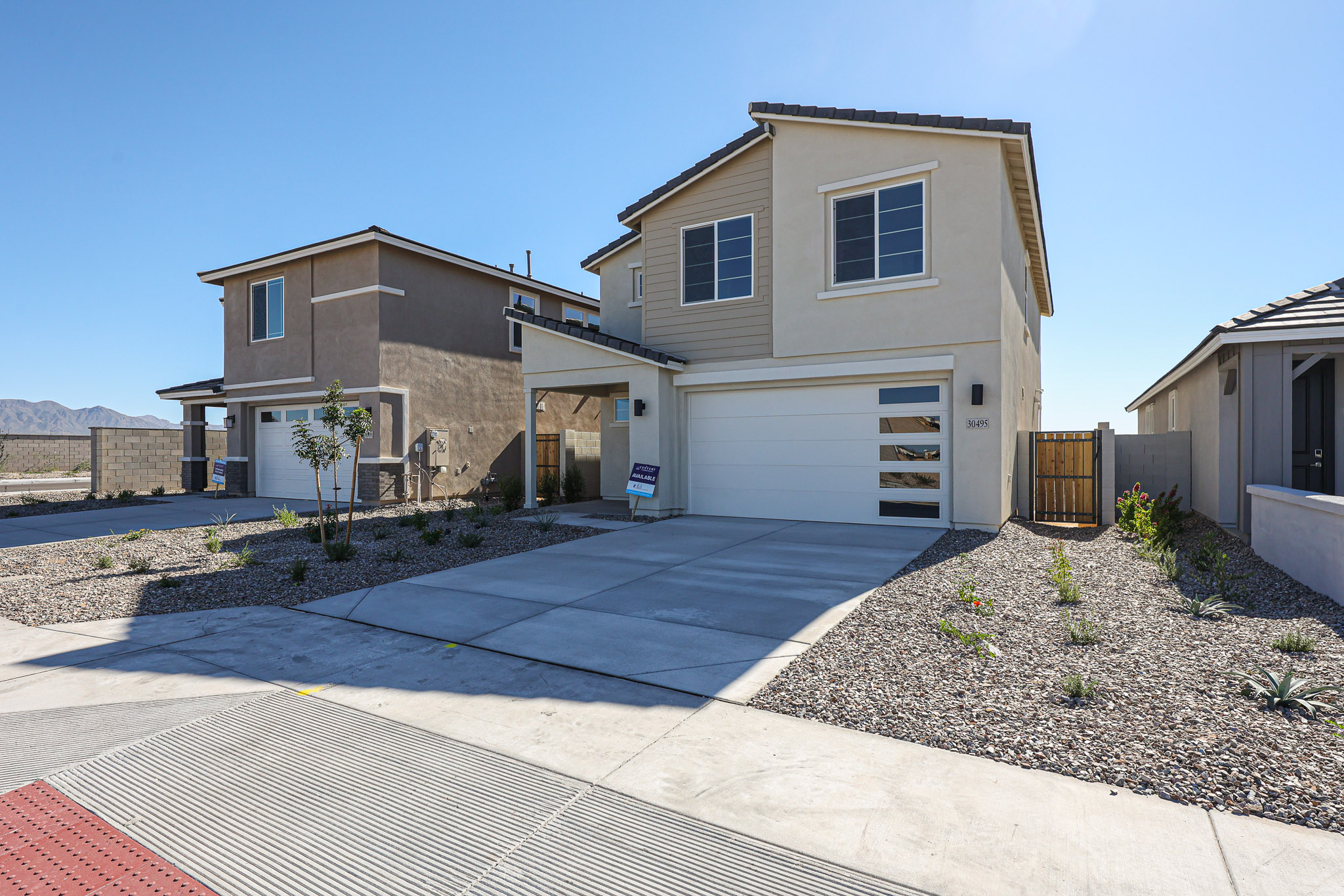 Two-story beige house with a white garage door, small front yard with gravel and desert plants, and a neighboring house on the left under a clear blue sky.
