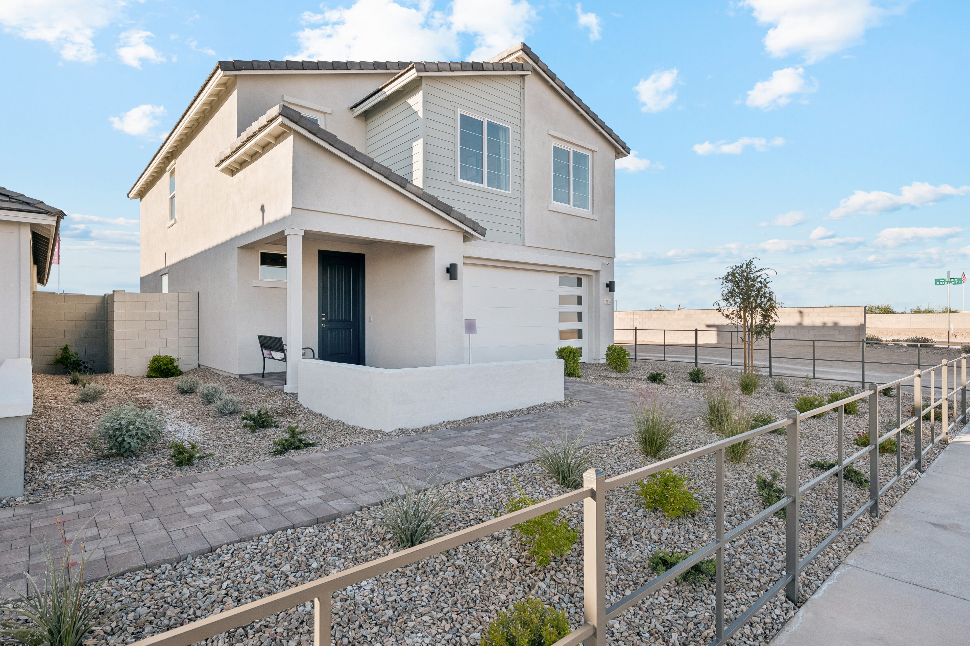 Two-story modern house with light-colored exterior, single-car garage, small front porch, and desert landscaping with gravel and minimal plants. A paved walkway leads to the front door.