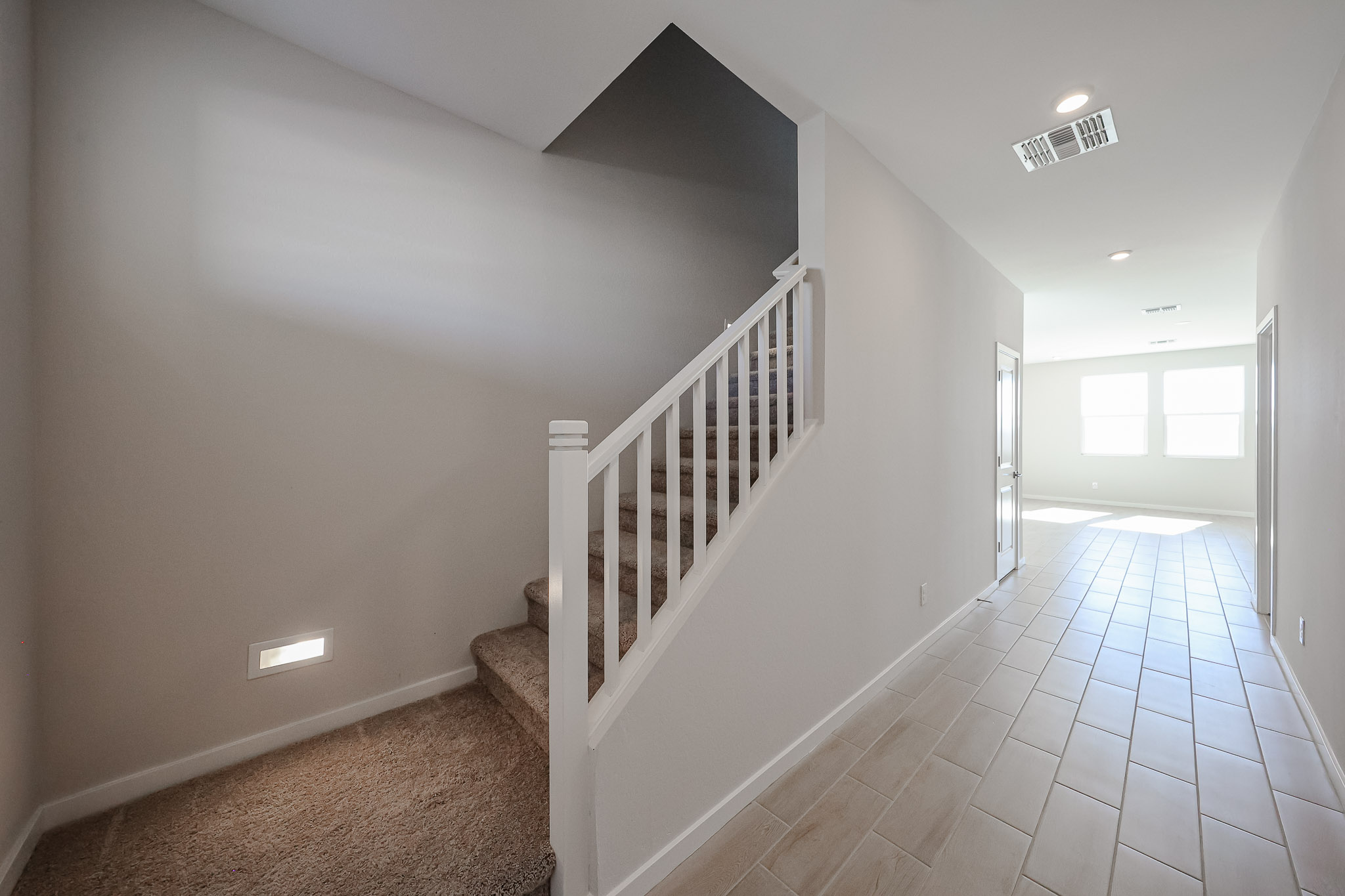 Interior hallway with a carpeted staircase on the left and a tiled floor leading to a bright room with large windows at the end.