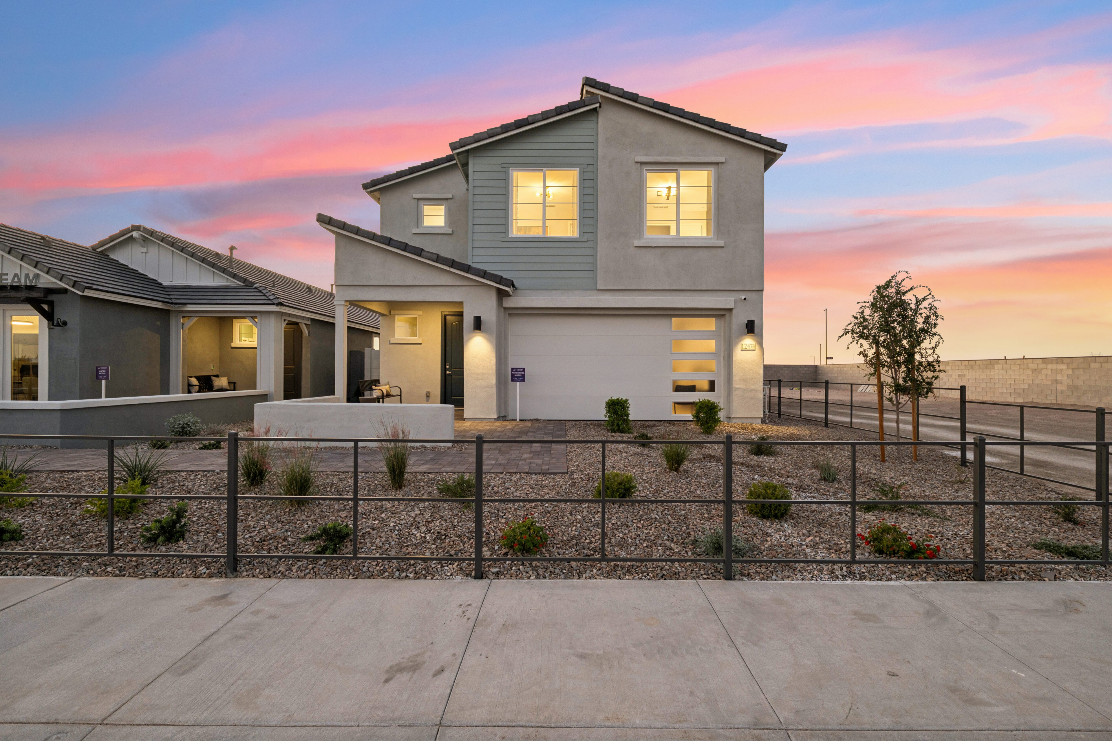 Modern two-story house with gray exterior, large windows, garage door, and xeriscaped front yard at sunset.