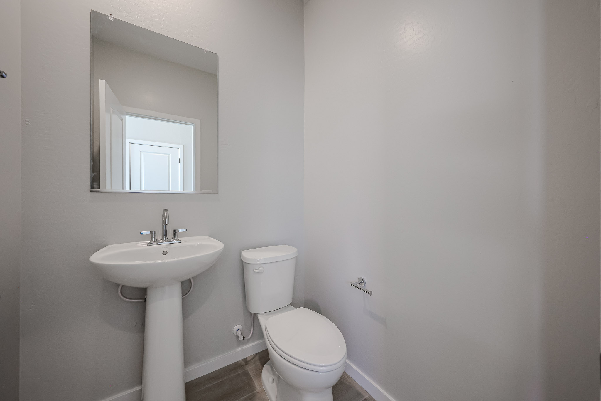 Small bathroom with a pedestal sink, wall-mounted mirror, toilet, toilet paper holder, and light gray walls.