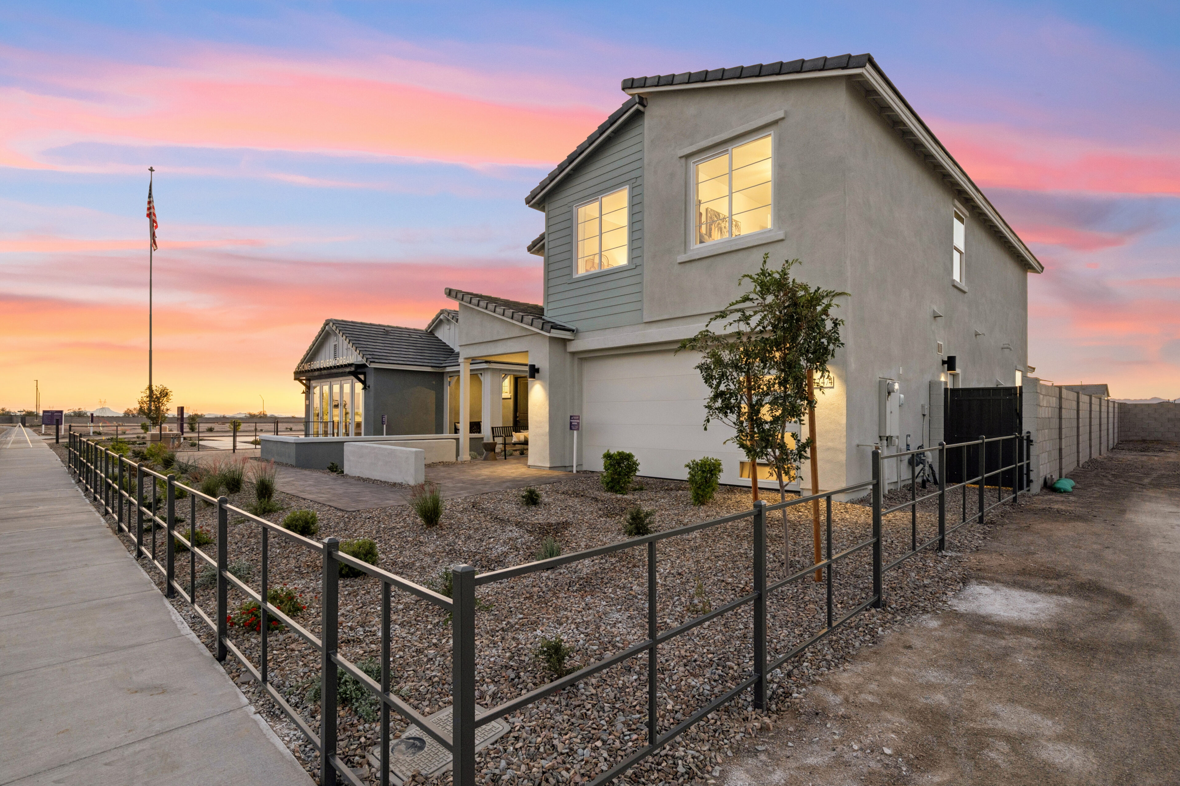 Modern two-story house with gray exterior, large windows, and a gravel yard, photographed at sunset. A flagpole stands nearby, and the property is enclosed by a metal fence.