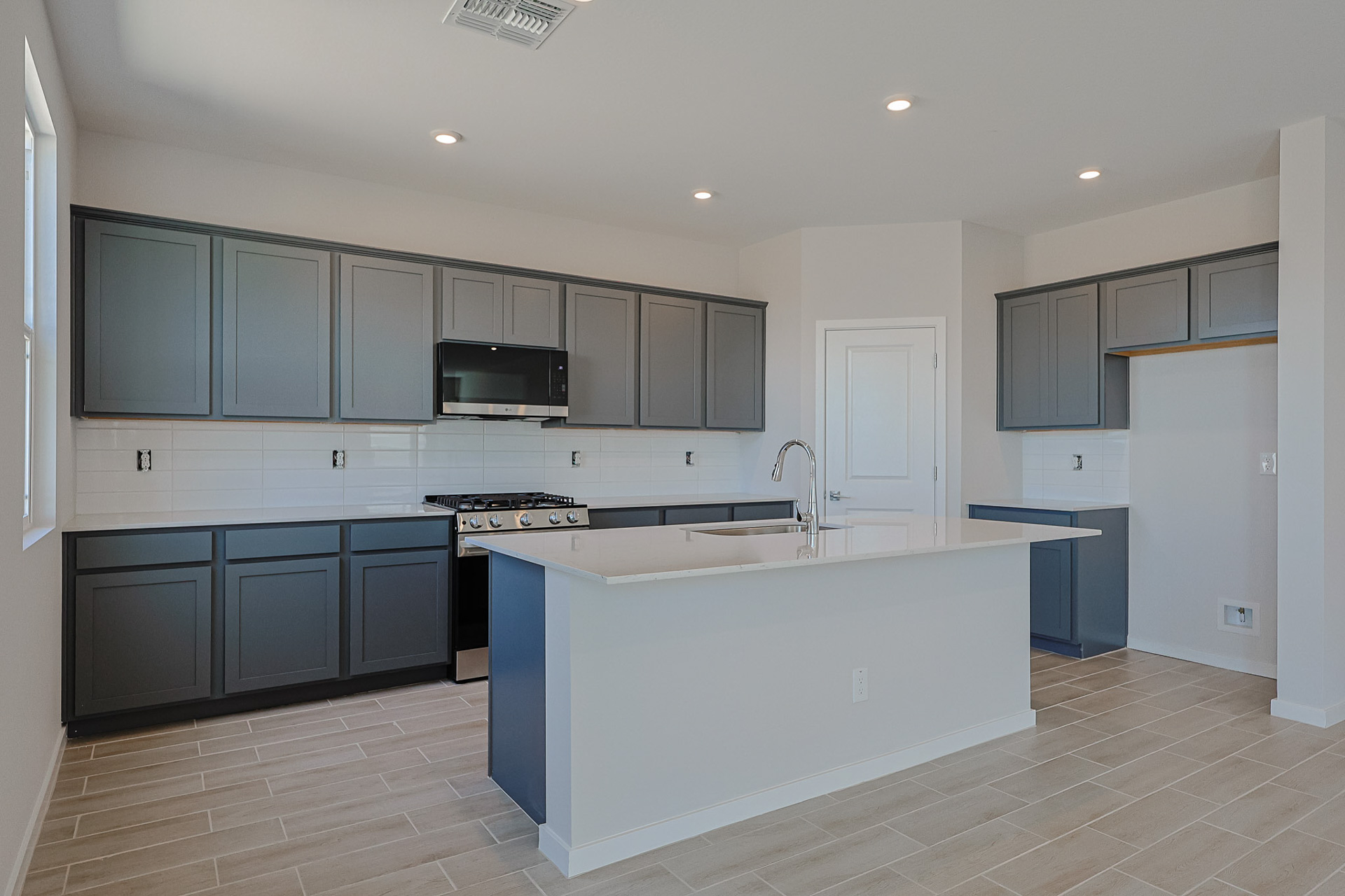 Modern kitchen with gray cabinets, stainless steel appliances, white tiled backsplash, center island with sink, and light-colored tile flooring.