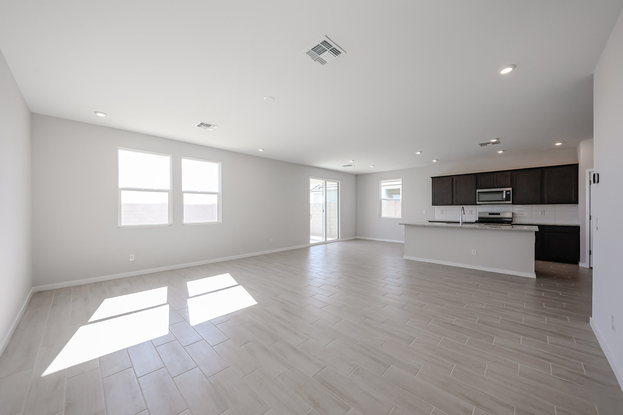 Spacious, empty open-plan living area with light tile flooring, white walls, and a modern kitchen with dark cabinets and stainless steel appliances. Large windows and glass doors provide natural light.