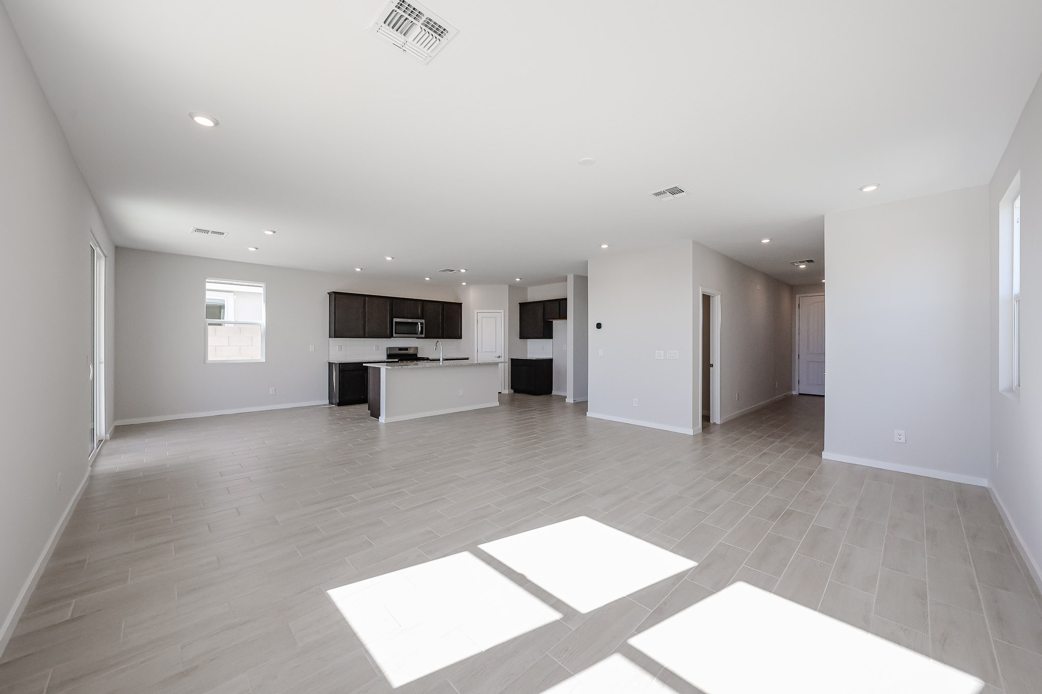 Spacious, unfurnished open-plan living area with light tile floors, recessed lighting, and a modern kitchen featuring dark cabinets and stainless steel appliances. Natural light from multiple windows.