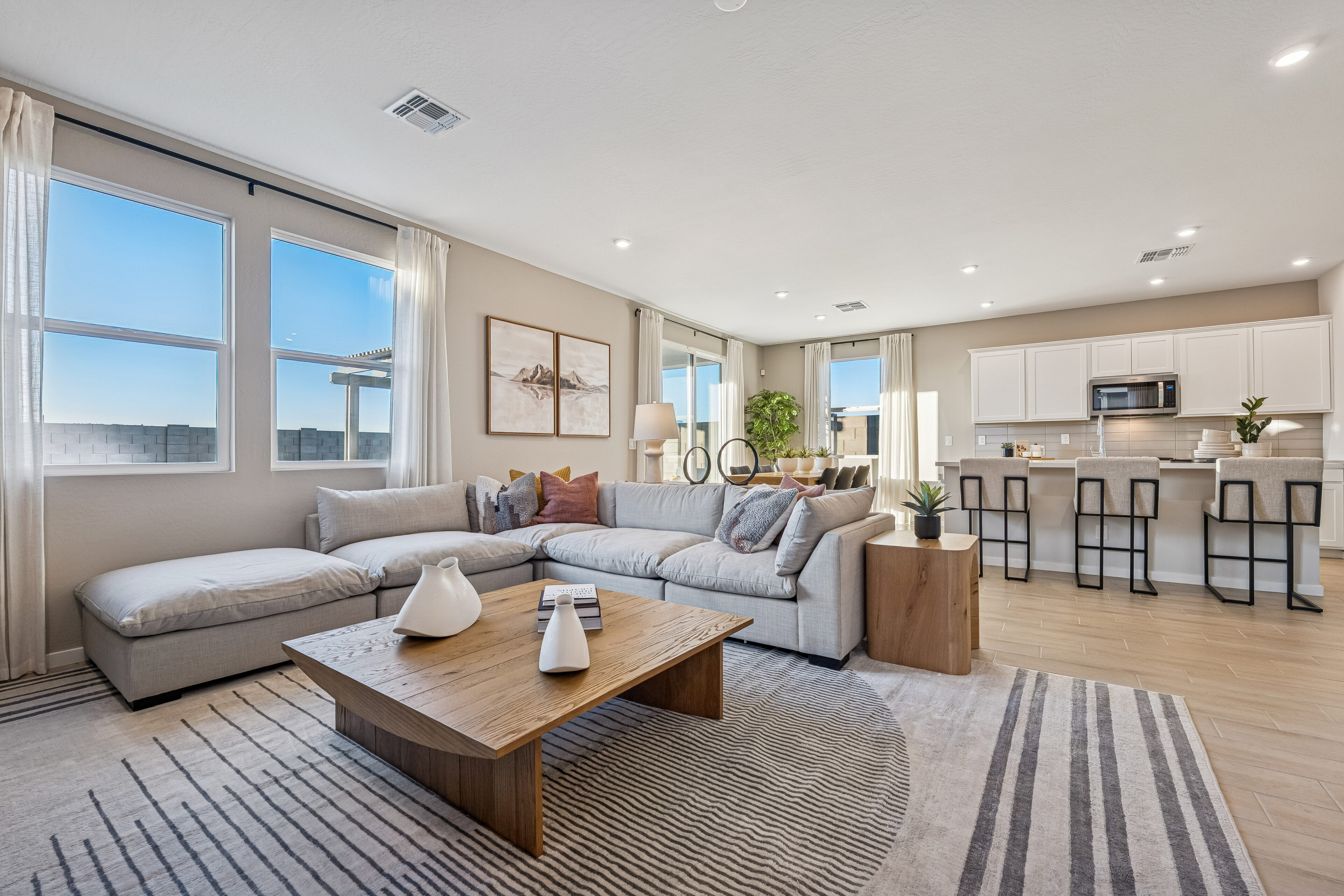 Modern open-concept living room and kitchen with neutral decor, large sectional sofa, coffee table, bar stools, and bright natural light from multiple windows.