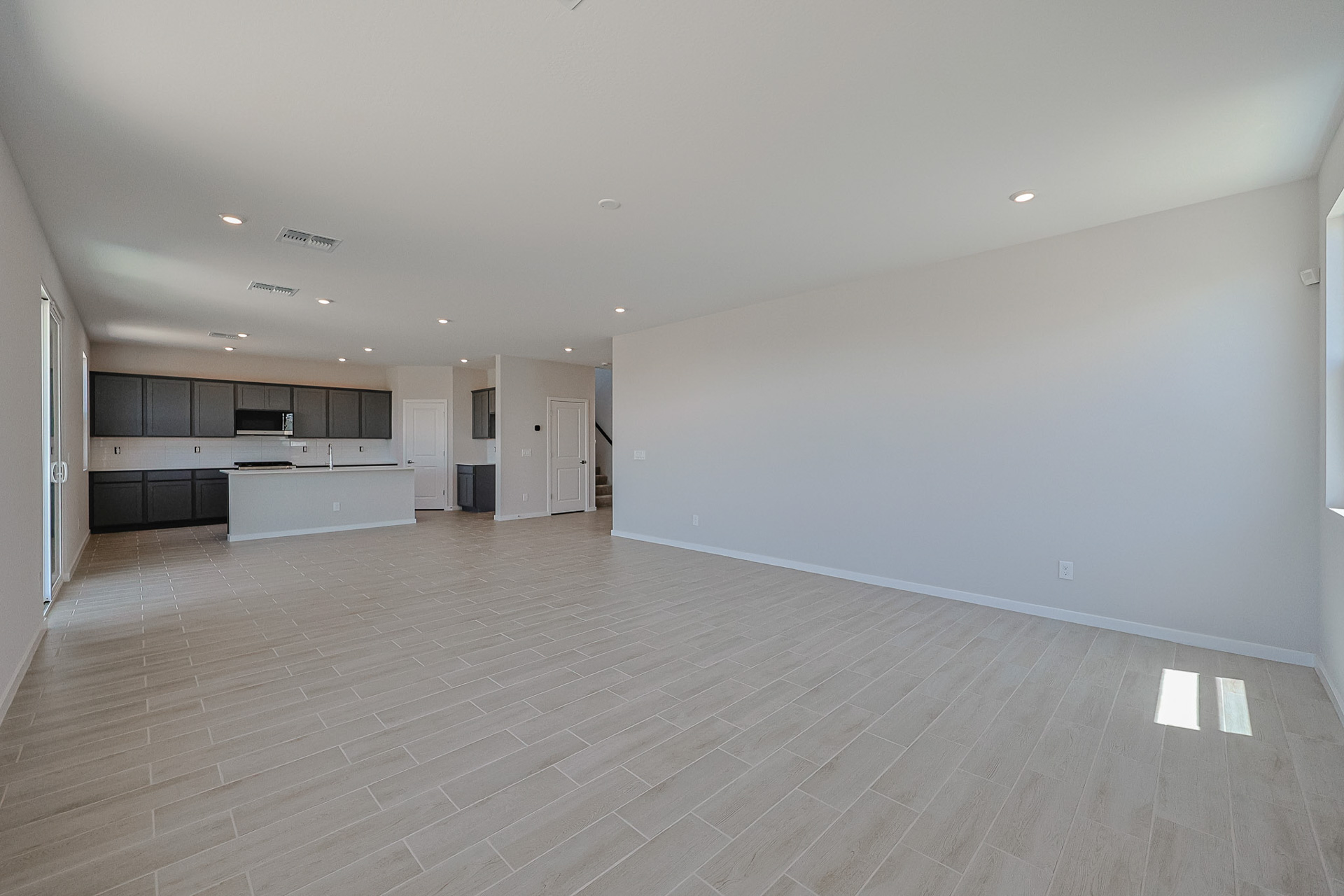 Spacious empty living area with light tile flooring, recessed lighting, and an open kitchen with dark cabinets in the background.