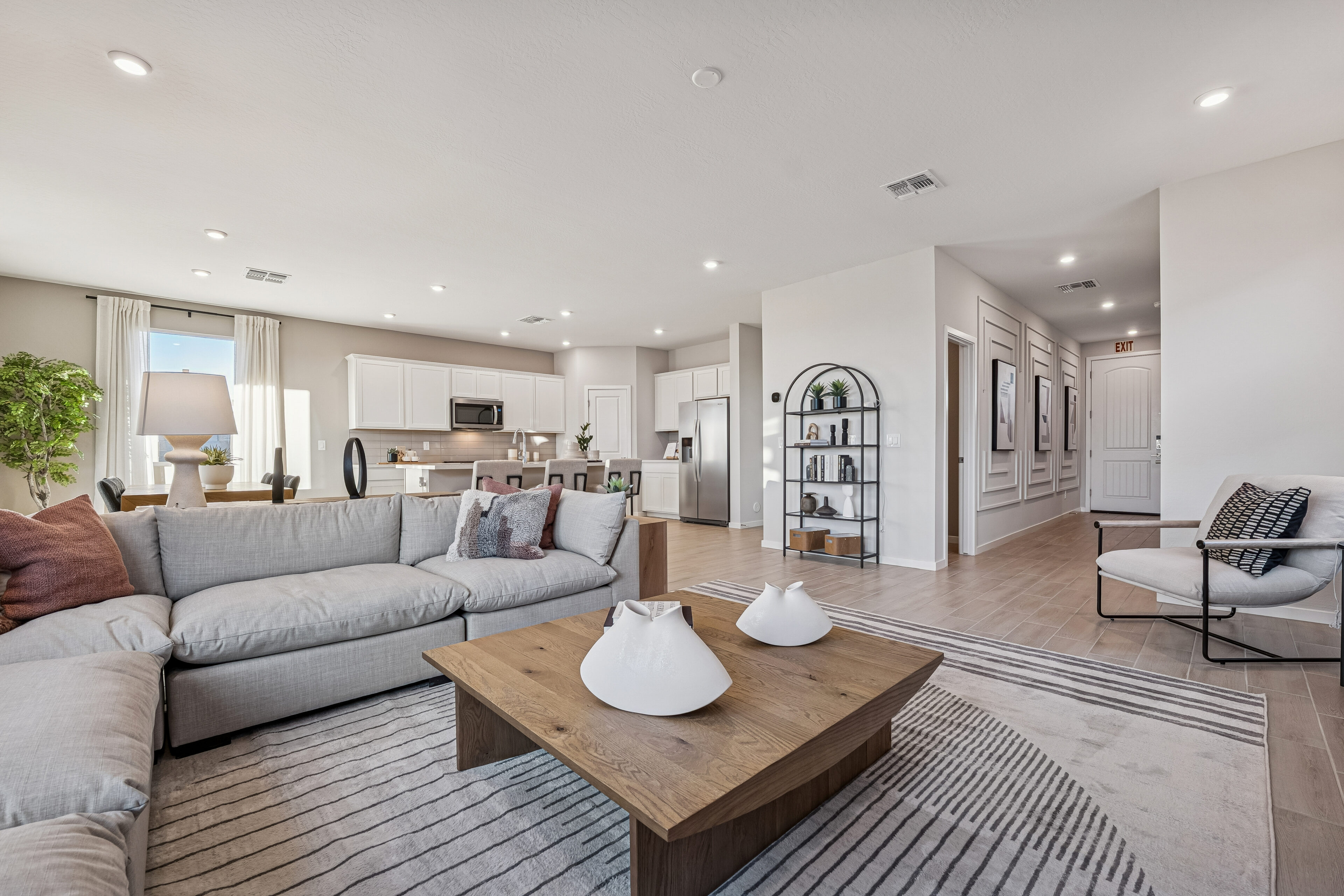 Modern open-plan living room and kitchen with neutral colors, a sectional sofa, wooden coffee table, striped rug, and minimalist decor. Large windows provide natural light.