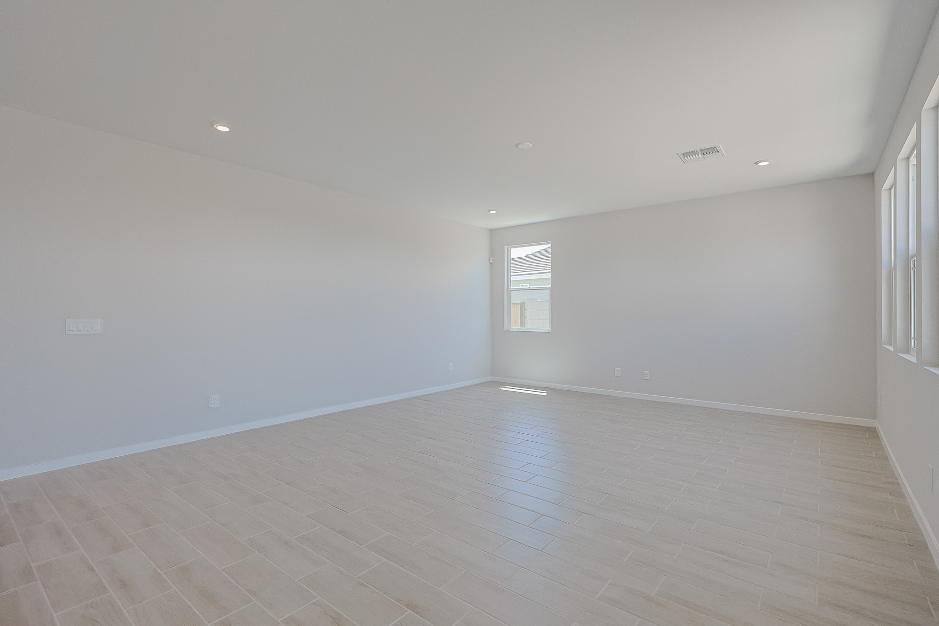 Spacious, empty room with light wood-look tile flooring, white walls, recessed lighting, and windows letting in natural light.