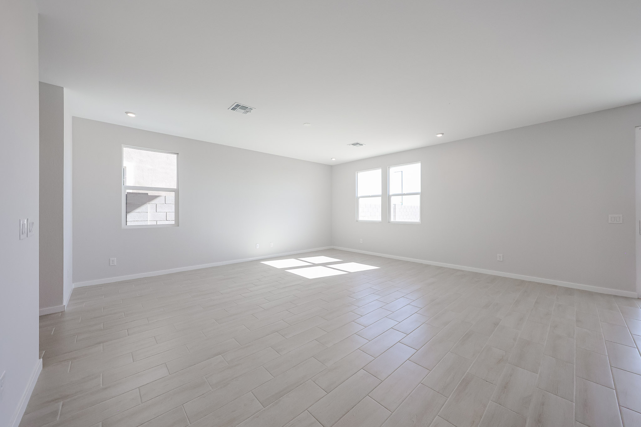 Empty, spacious room with light-colored tile flooring, white walls, and multiple windows letting in natural light. Ceiling lights and air vents are visible.