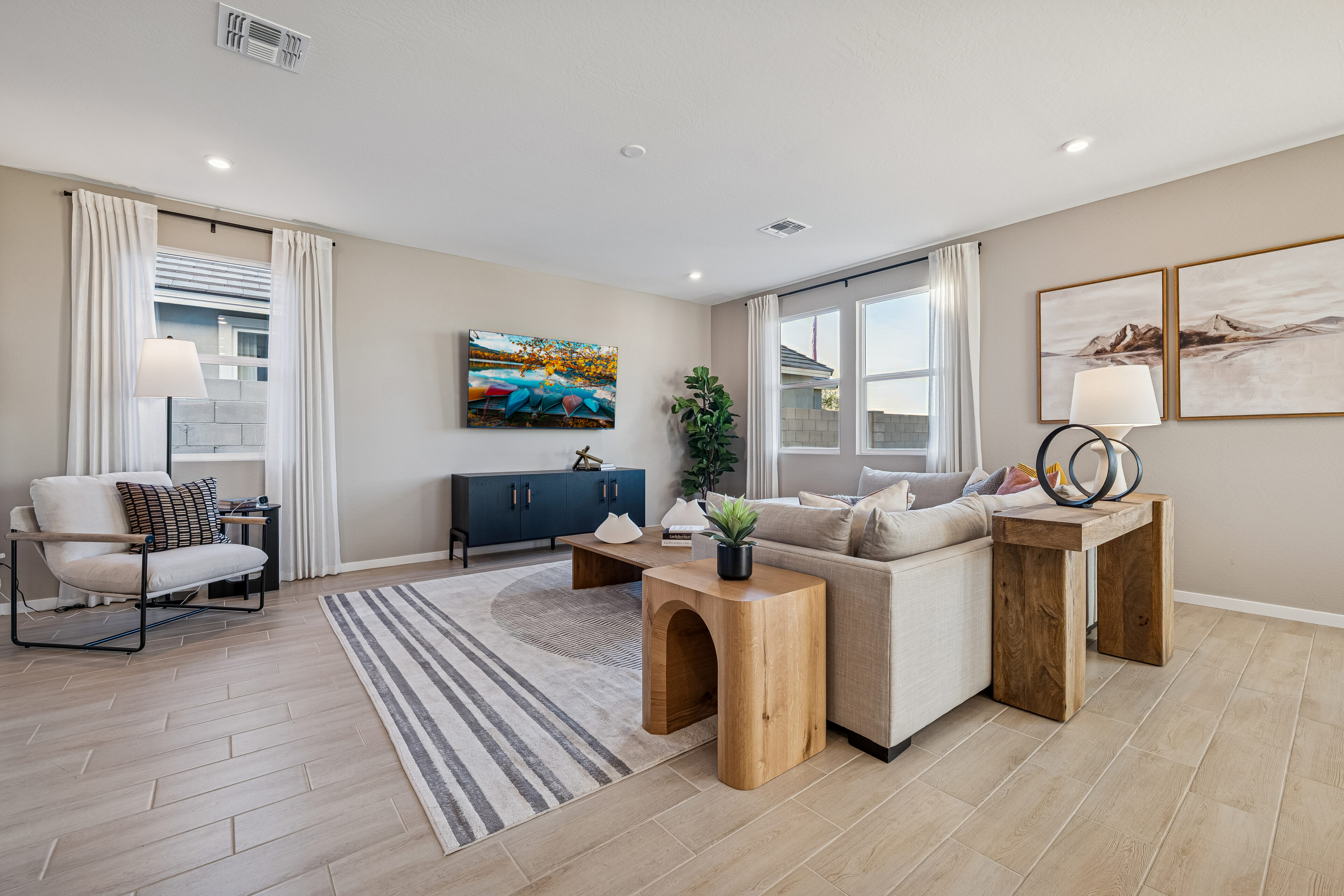 Modern living room with a beige sectional sofa, wooden tables, large windows, neutral decor, wall-mounted TV, and framed art on light-colored walls.