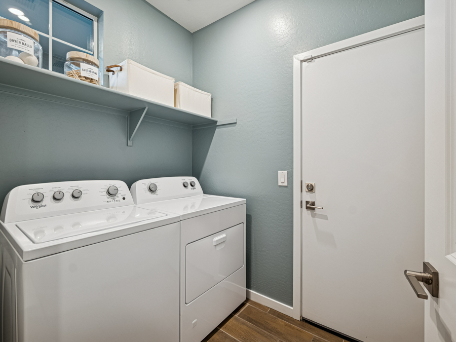 Laundry room with a white washing machine and dryer, a shelf above holding storage bins and detergent, blue walls, wood flooring, and a closed white door.