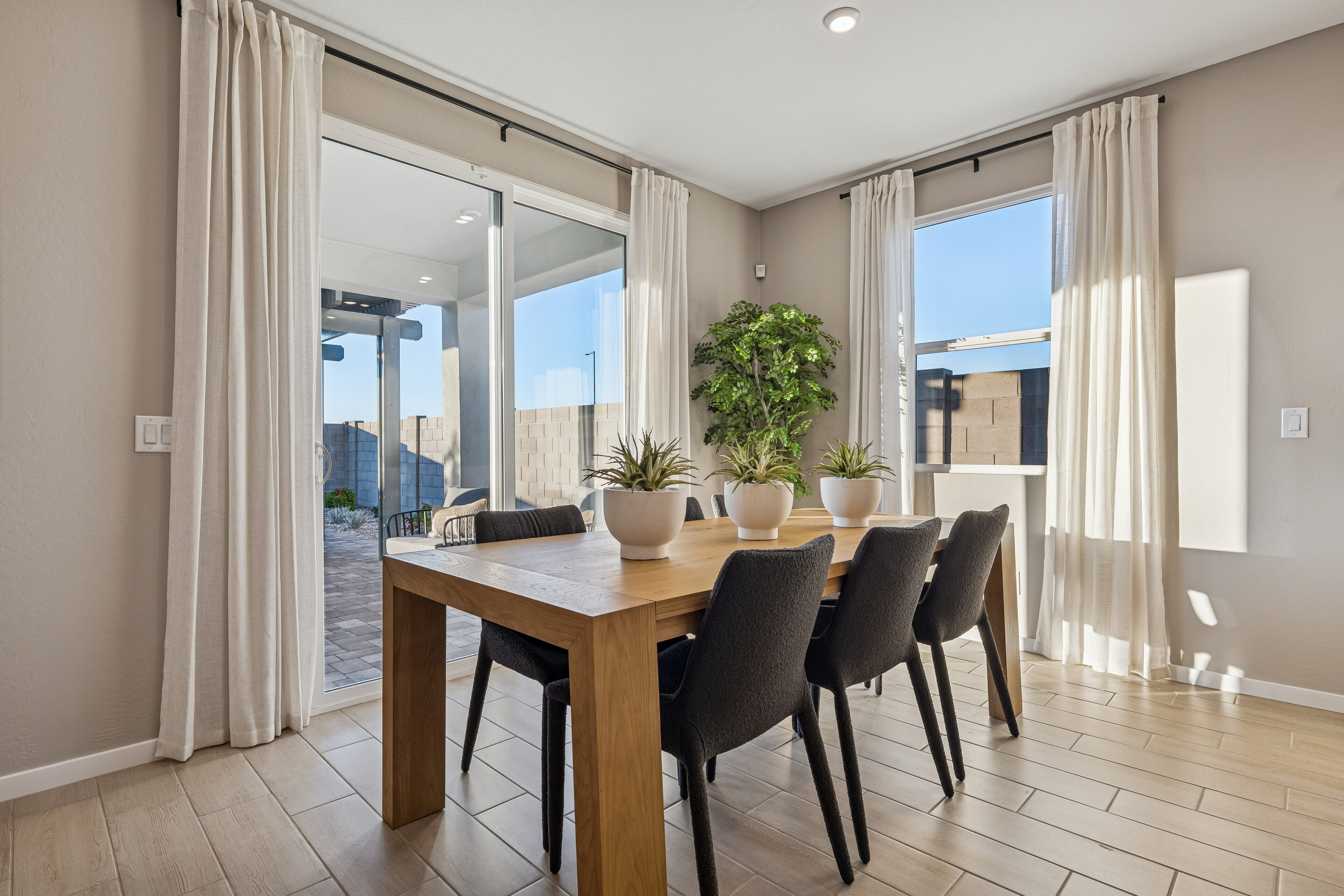 Modern dining room with a wooden table, six black chairs, potted plants, light curtains, tile flooring, and large windows showing an outdoor patio area.