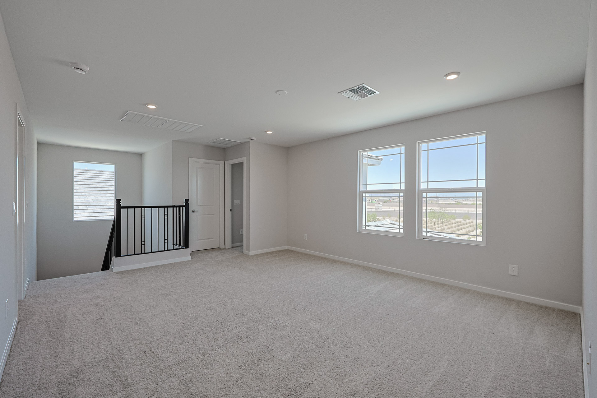 Unfurnished, carpeted upstairs room with white walls, two windows, ceiling lights, and a black railing by the staircase. Door and vent are on the left wall.