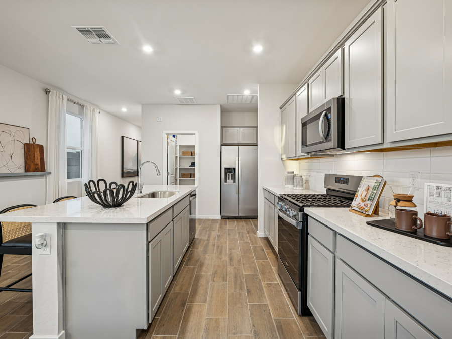 Modern kitchen with gray cabinets, stainless steel appliances, wood-look tile flooring, an island with a sink, and a walk-in pantry. Bright lighting and neutral decor complete the space.