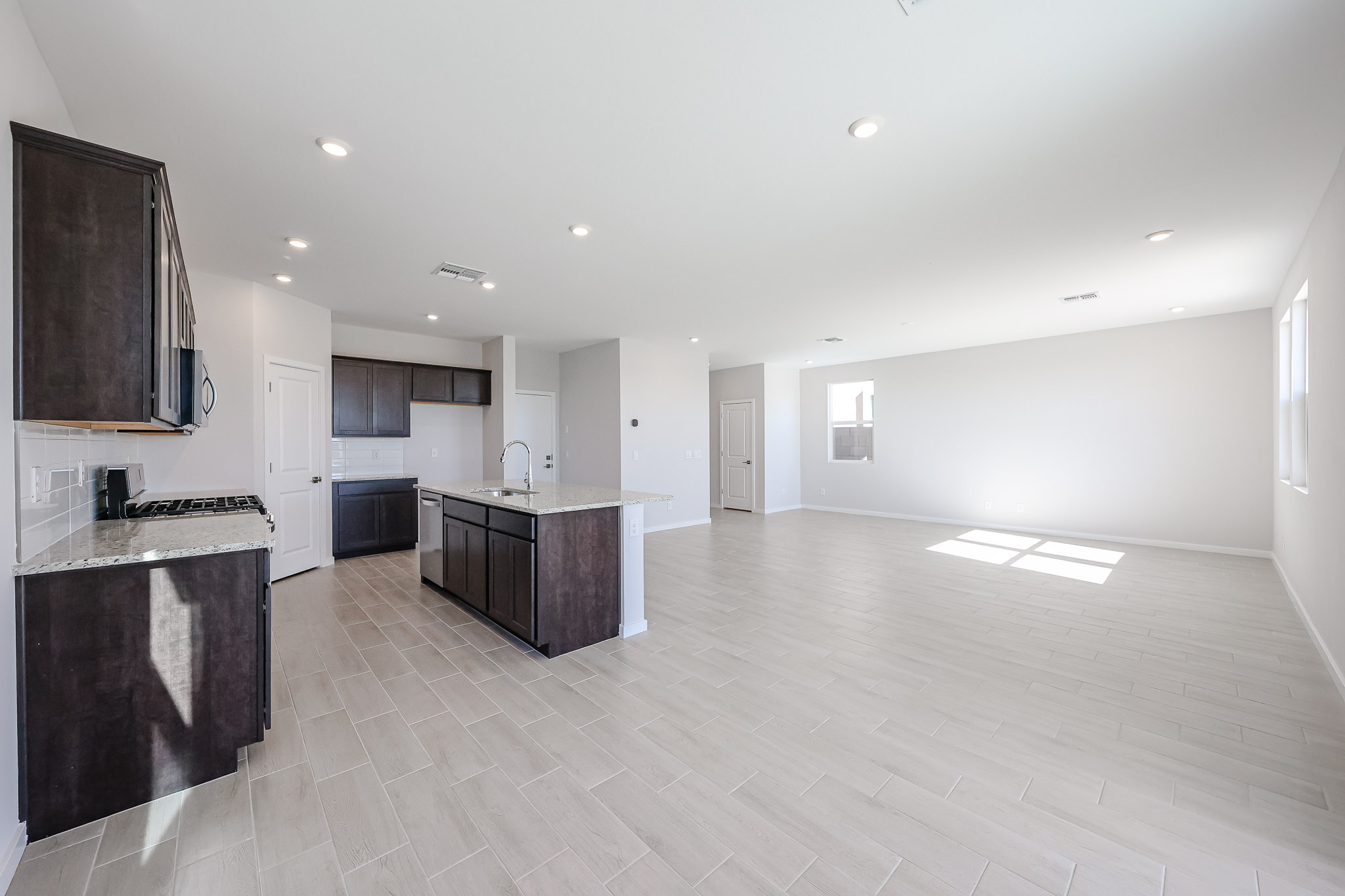 Spacious, empty open-plan kitchen and living area with light tile floors, white walls, dark cabinetry, and ample natural light from multiple windows.