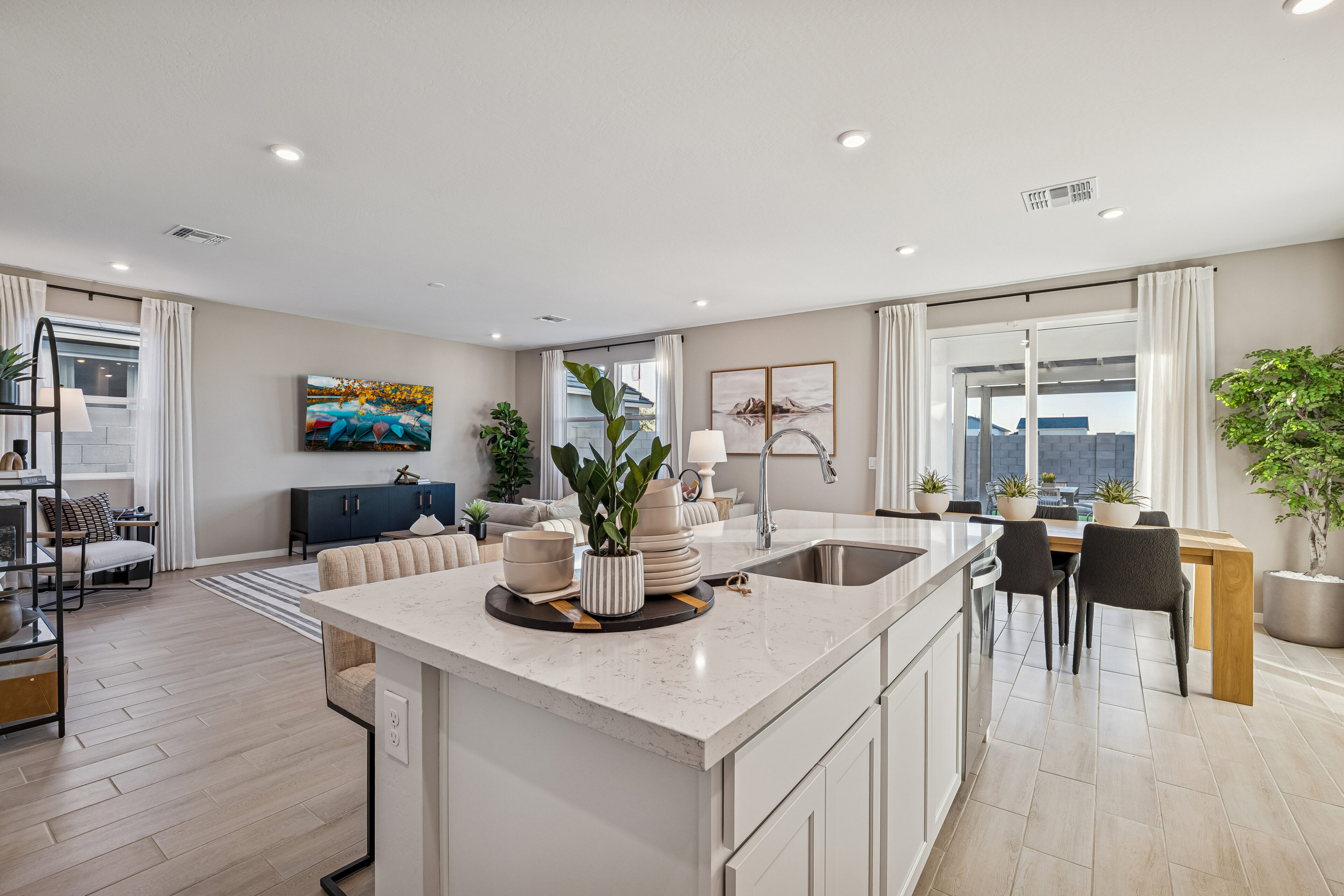 Modern open-concept kitchen and living area with island, dining table, sofa, TV, large windows, light wood floors, and potted plants.