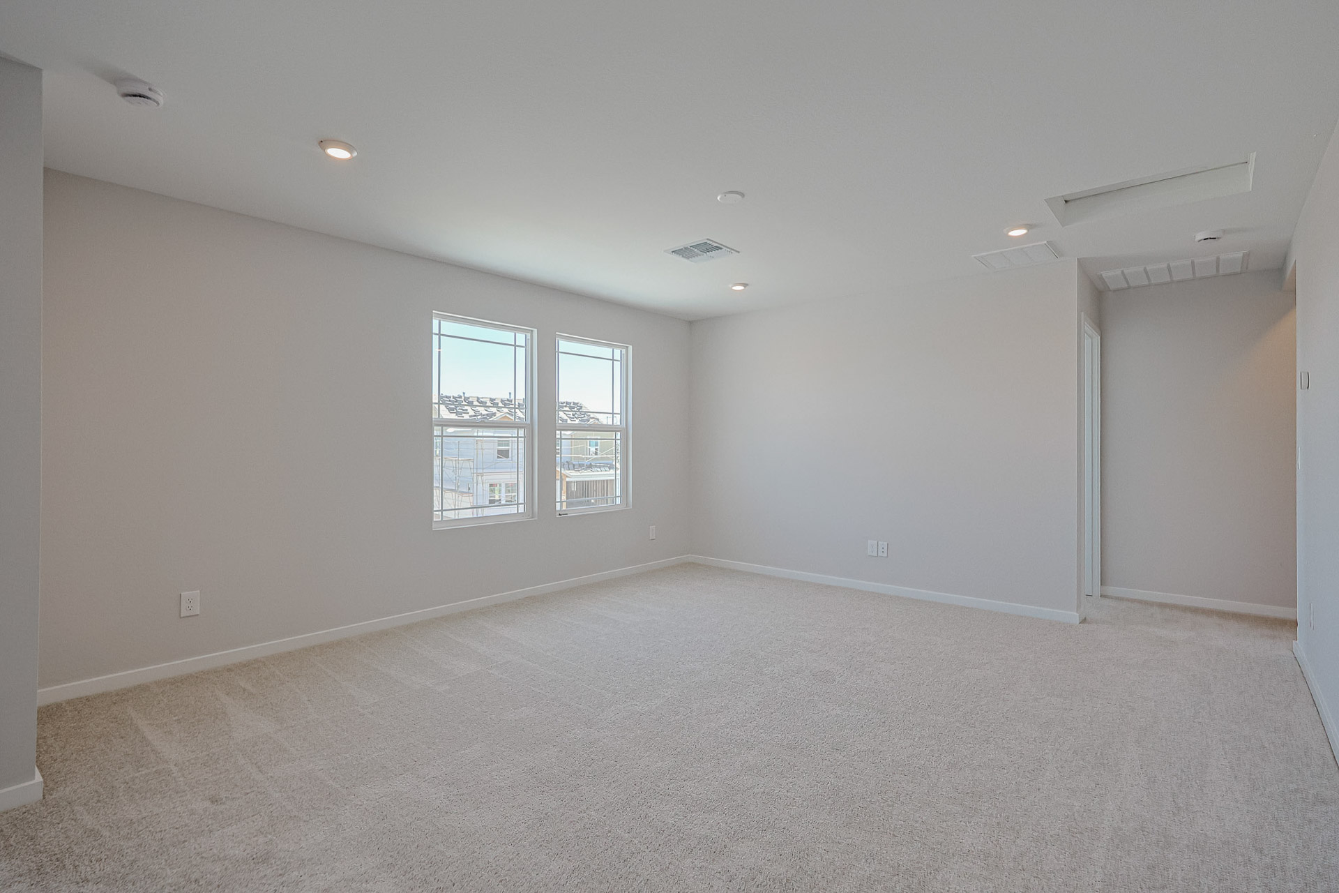 Empty carpeted room with two windows, recessed ceiling lights, and white walls; hallway entrance visible on the right.
