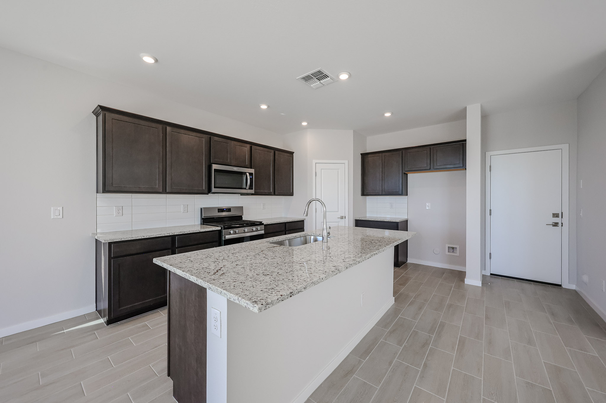 Modern kitchen with dark wood cabinets, granite countertops, stainless steel appliances, tile flooring, and white walls. Center island features a built-in sink and a chrome faucet.