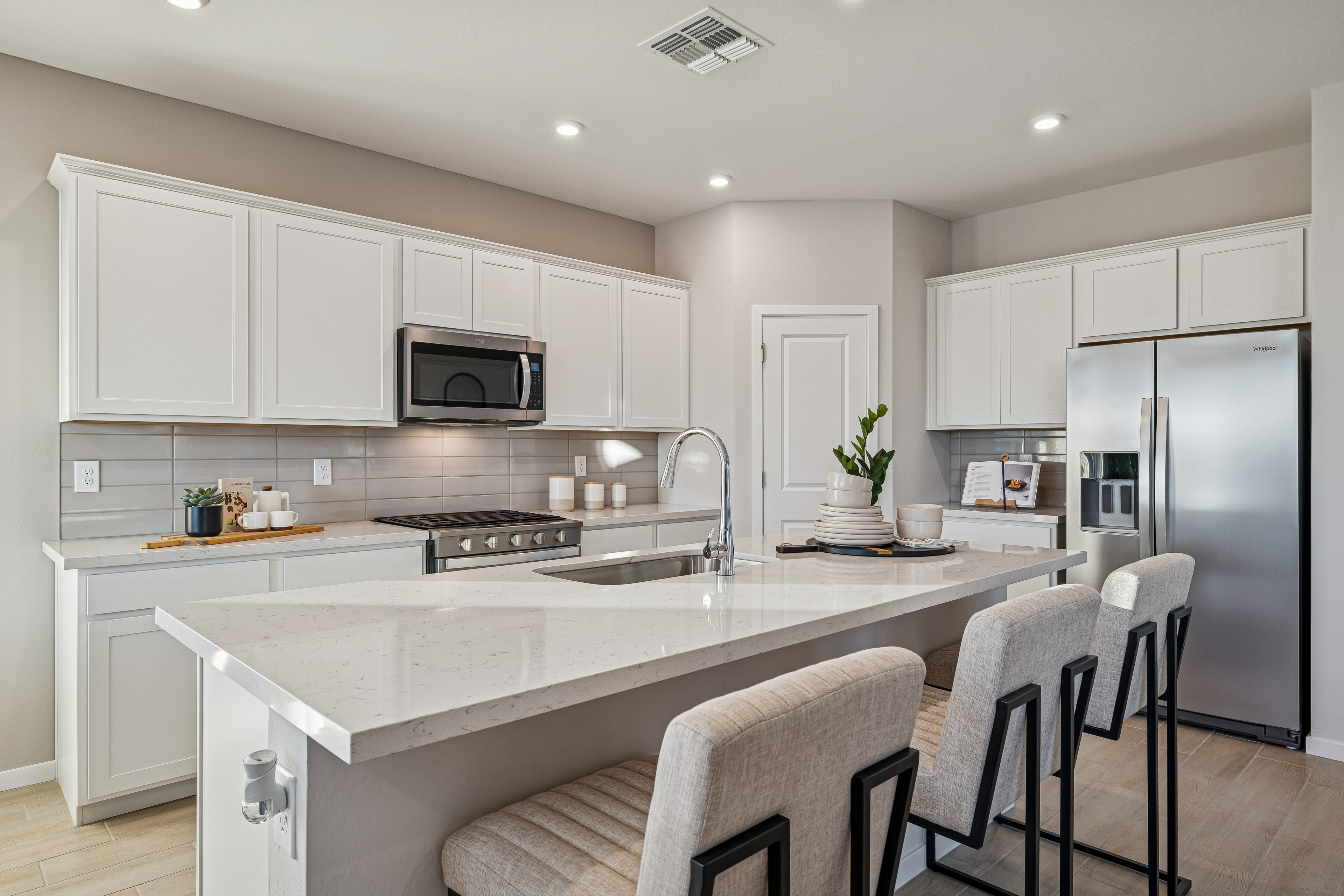 Modern kitchen with white cabinets, stainless steel appliances, an island with a sink, three upholstered barstools, and neutral decor. Bright, clean, and organized space.