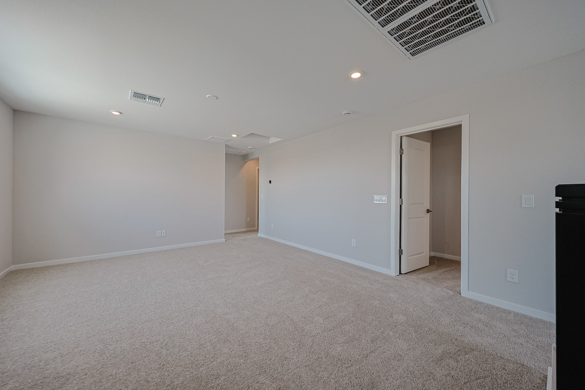 Empty carpeted room with light-colored walls, recessed lighting, ceiling vents, and an open door leading to another area. No furniture or decorations are present.