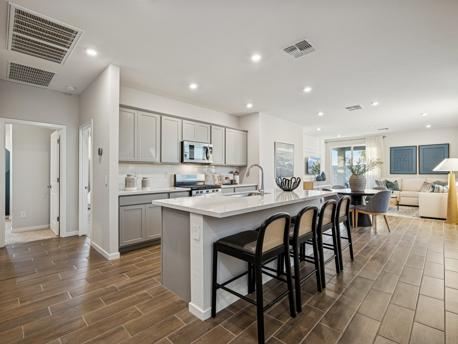 Modern kitchen with gray cabinets, white countertops, and a large island with three barstools; open layout connects to a living area with beige sofas and wood-look tile flooring throughout.