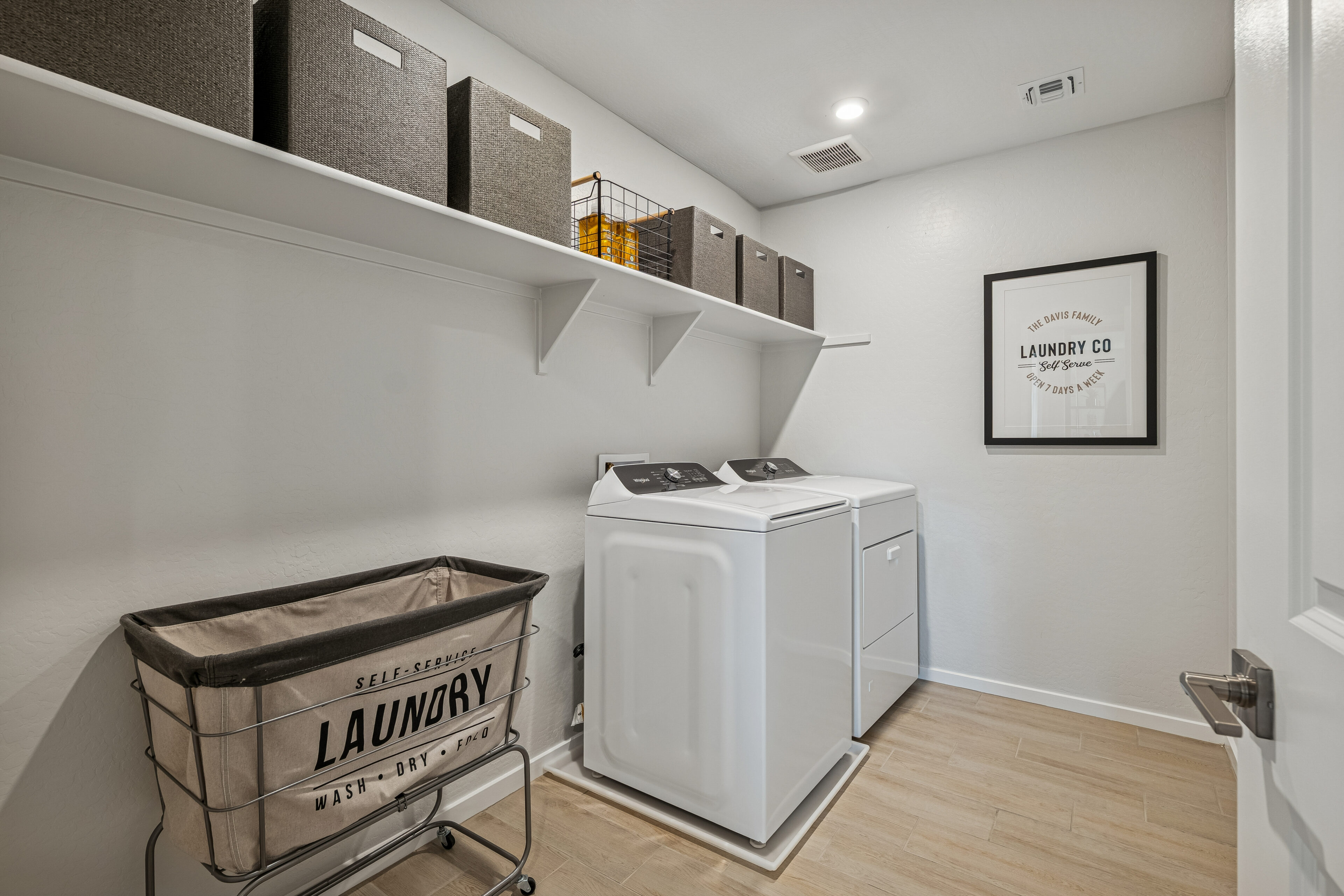 Modern laundry room with washer, dryer, rolling laundry hamper, upper shelf holding storage bins, and framed wall art on white walls.