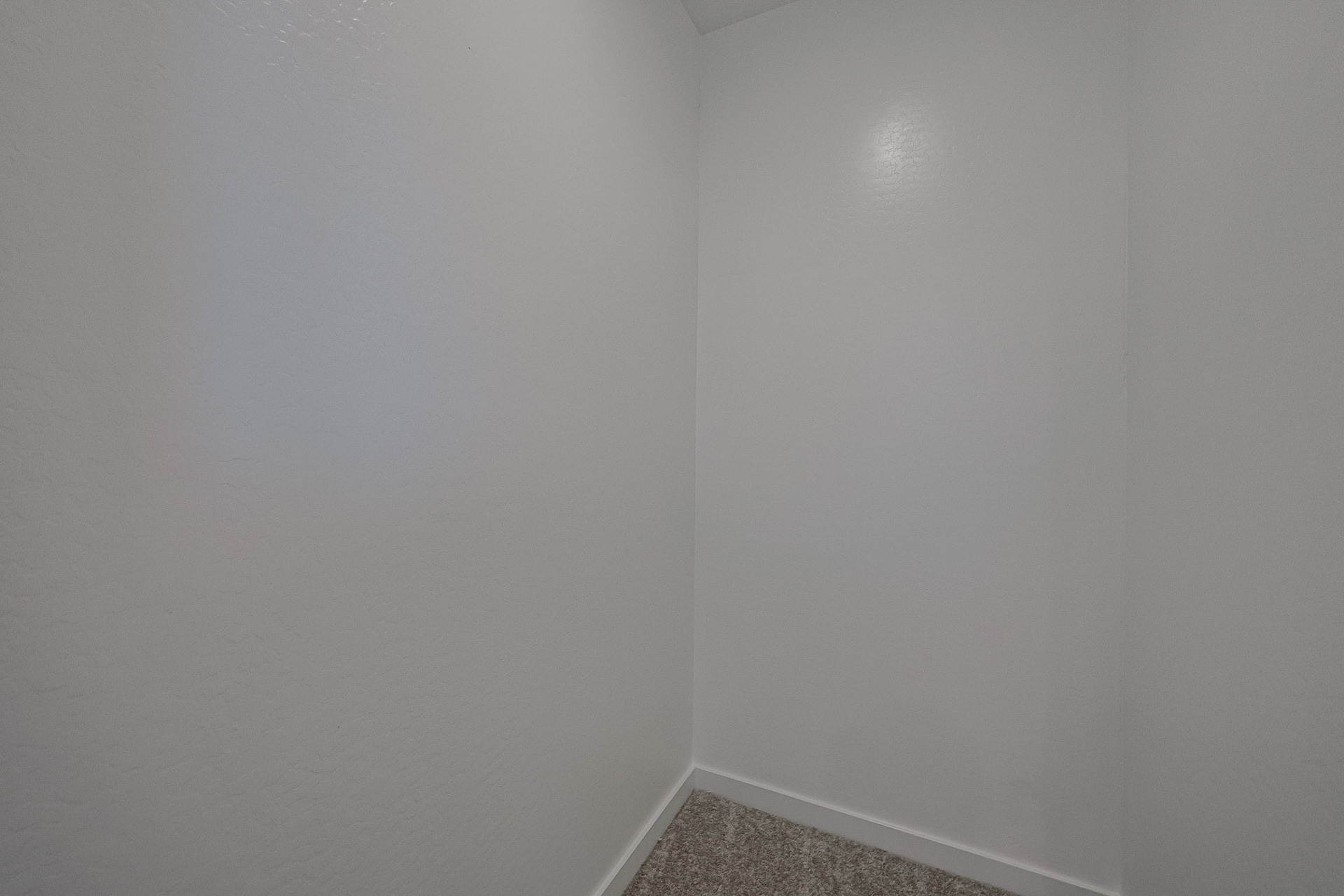 Empty, small corner room with plain white textured walls and a section of brown carpeted floor visible at the bottom.
